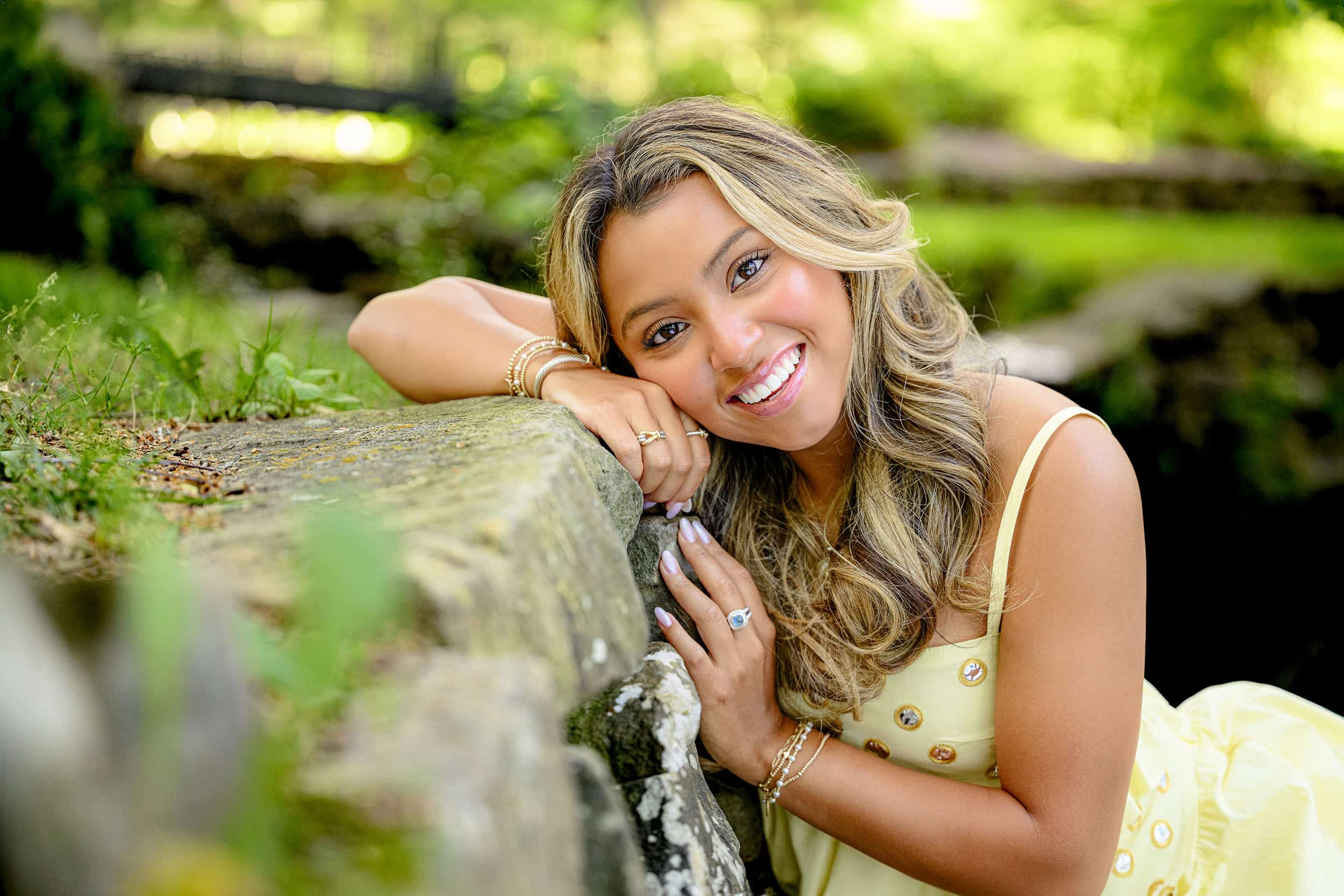 Blonde Charlotte Catholic Senior leans against a rock smiling with her hand under her cheek during her grad photos