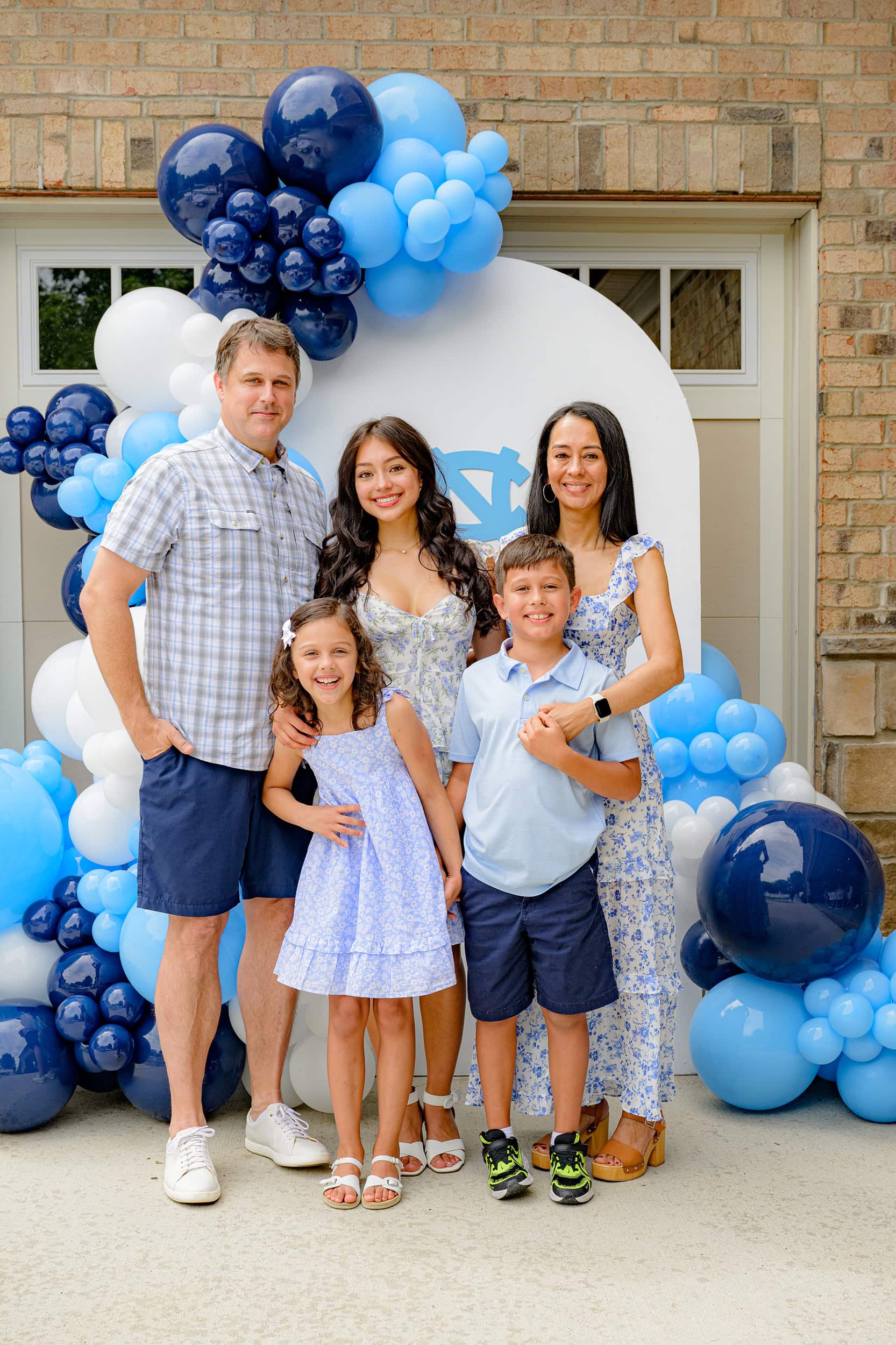 Family of 5 stands in front of a balloon arch decoration for a professional photo all dressed in the future graduates college colors of blue and white
