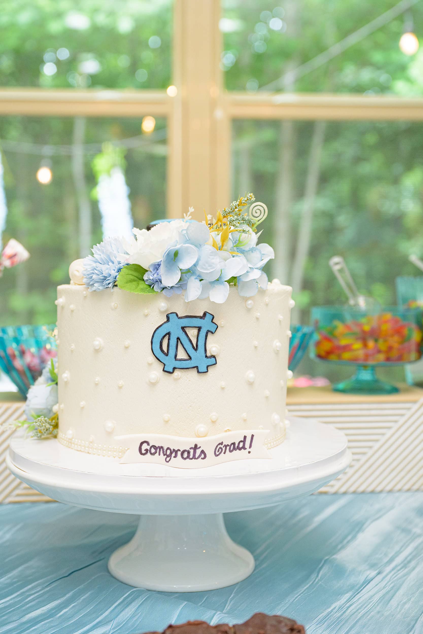 UNC CHapel Hill themed cake with logo, pearls, and congrats grad banner on it topped with blue toned flowers