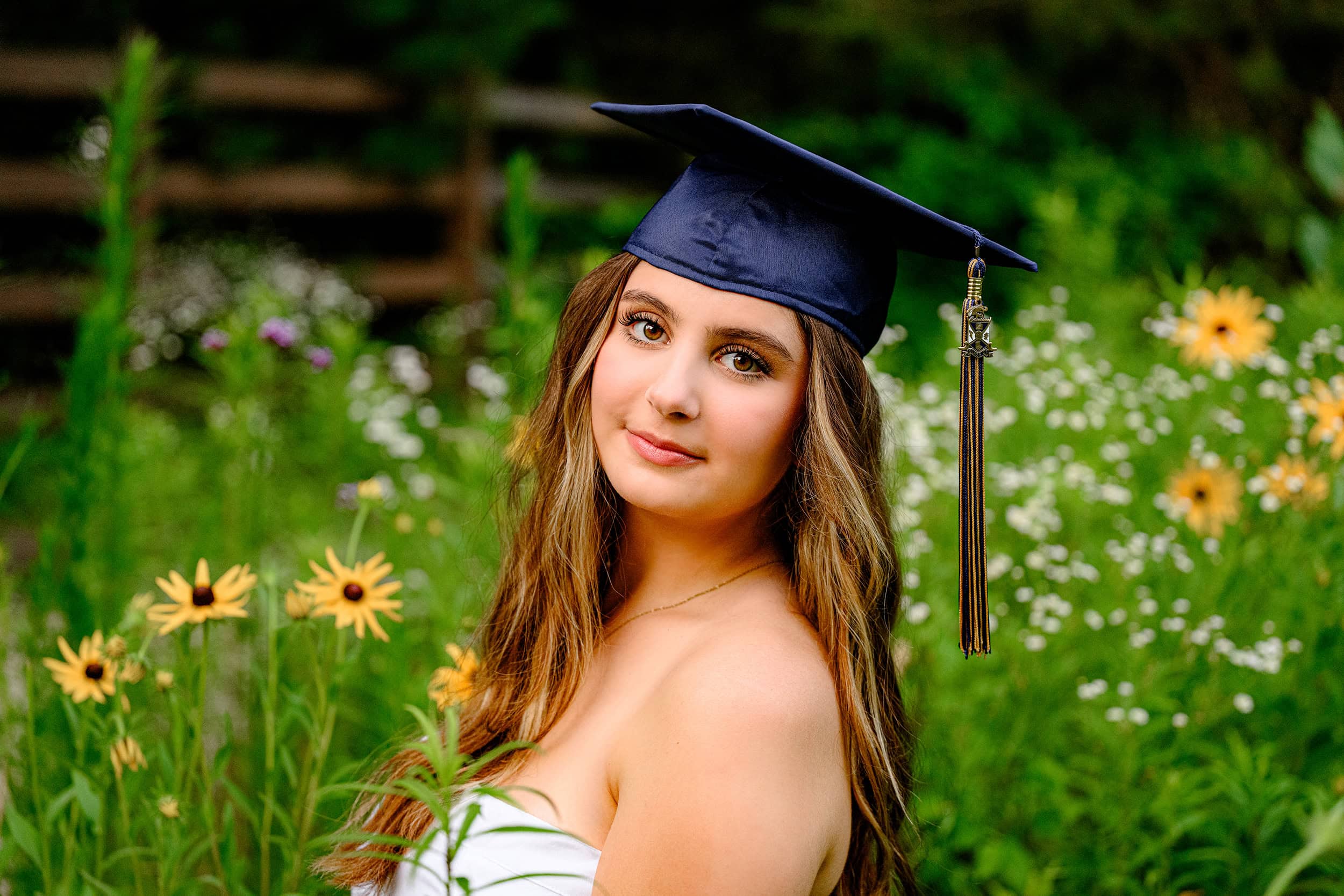 Cuthbertson high school graduate sits in flower garden wearing white graduation dress and her cap and tassel during her senior portraits