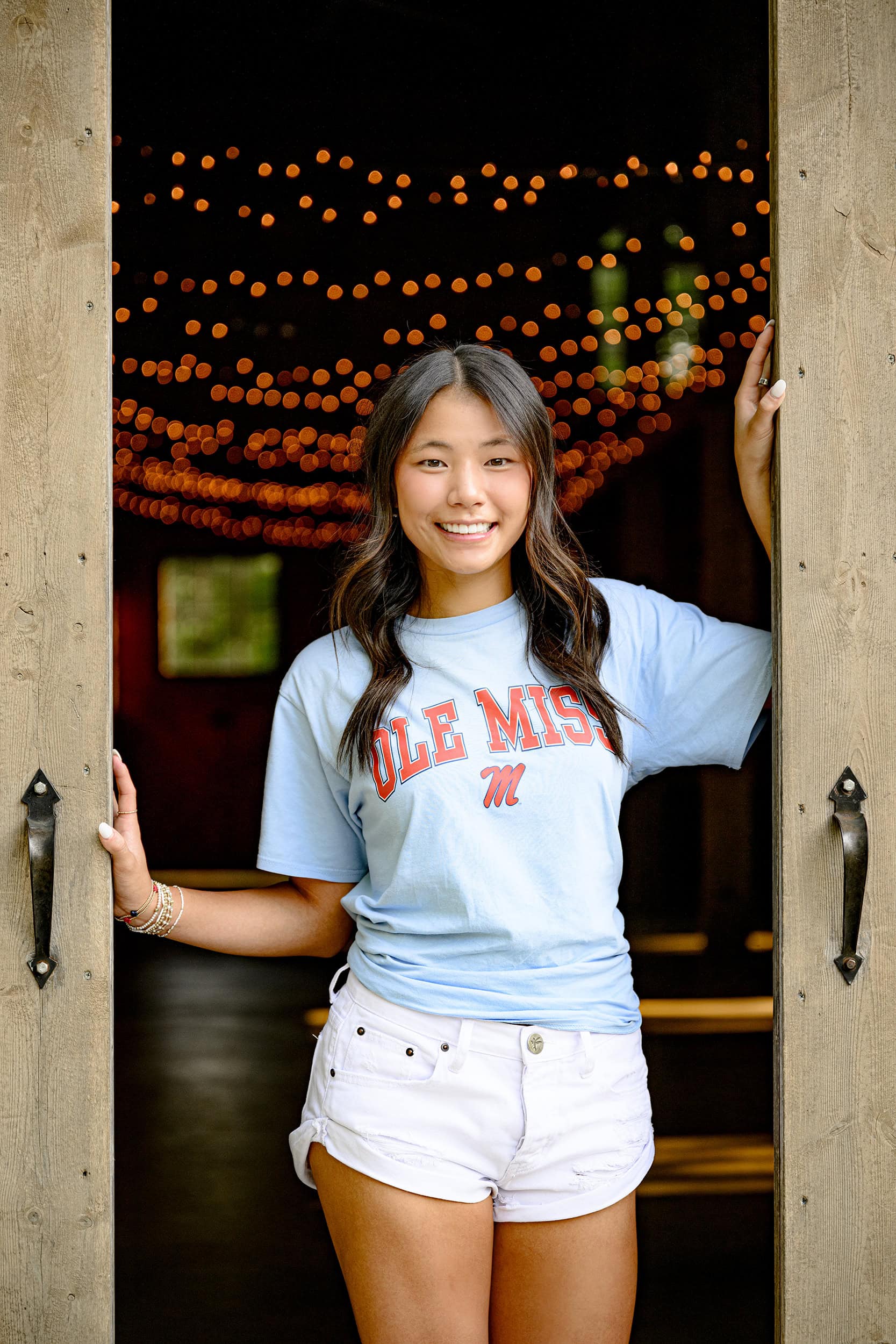 barn doors frame this Charlotte Catholic senior as she stands in her Ole Miss Tee shirt and shorts celebrating her next adventure themed senior photo session