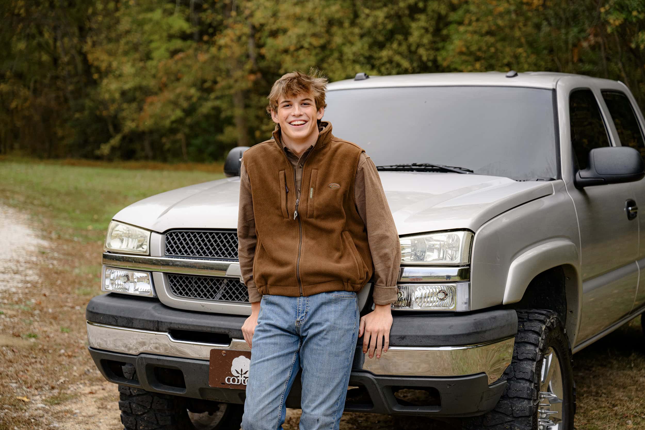 boy from indian land sc high school wearing brown shirt and vest leans against his silver truck in a fall field for his senior photos