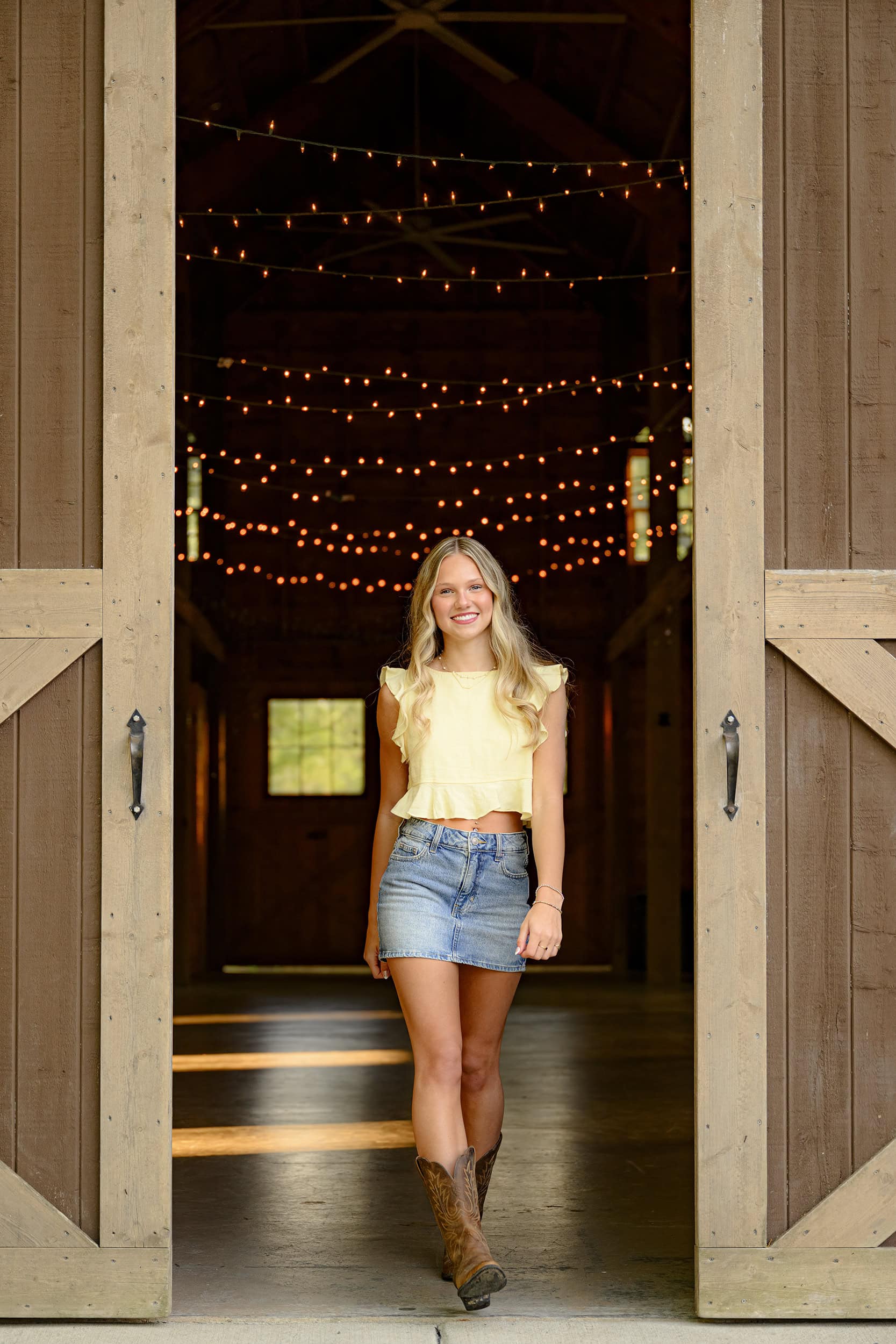 blonde parkwood high school senior wears yellow crop top and jean skirt with brown cowboy boots in a barn entrance with twinkle lights behind her in charlotte nc