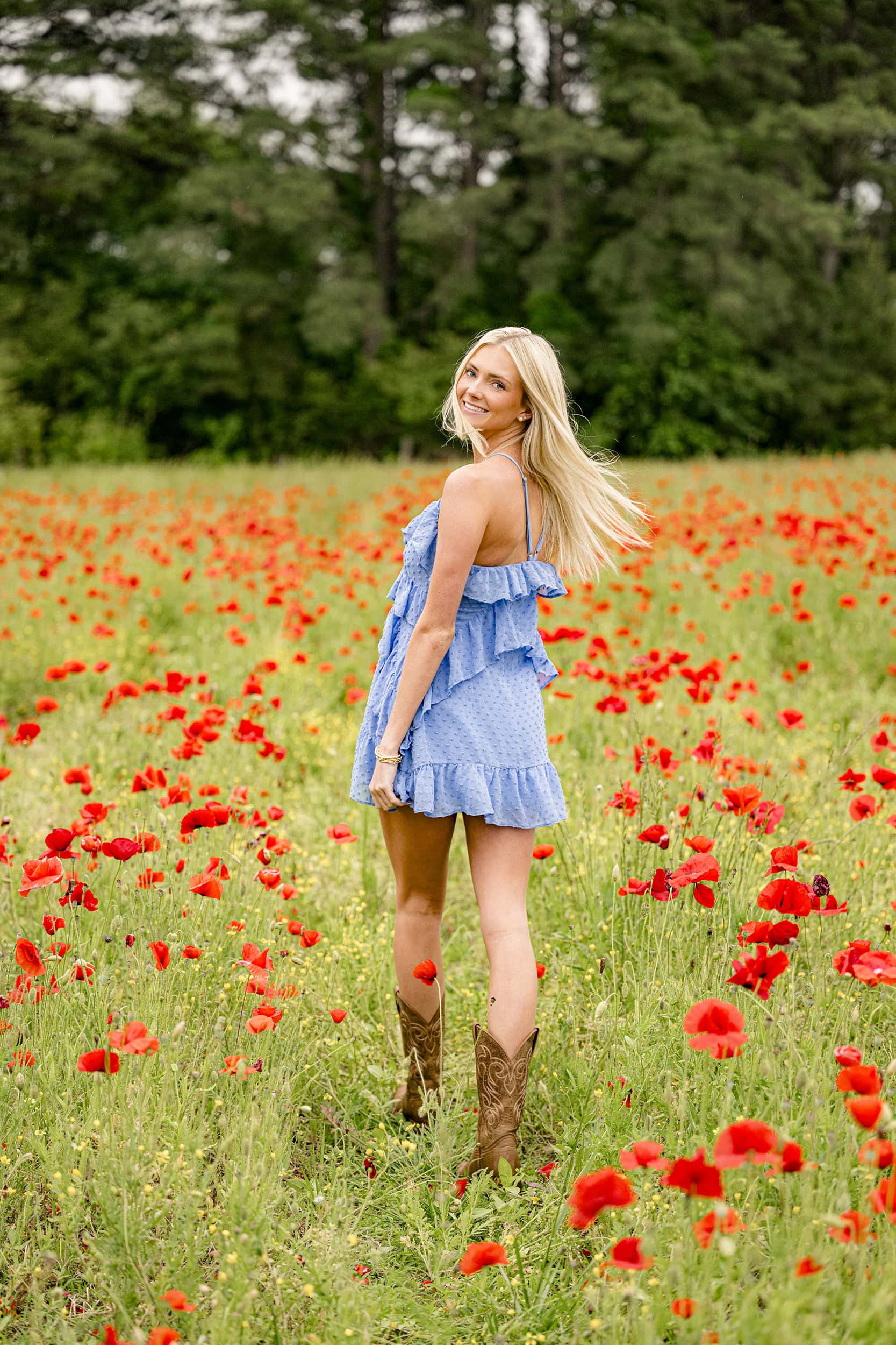 Blonde Cuthbertson high school soccer player K. Poole wearing blue dress and brown cowboy boots walks in a field of red poppies at Blue Hill Gardens in Monroe, NC with her hair blowing behind her