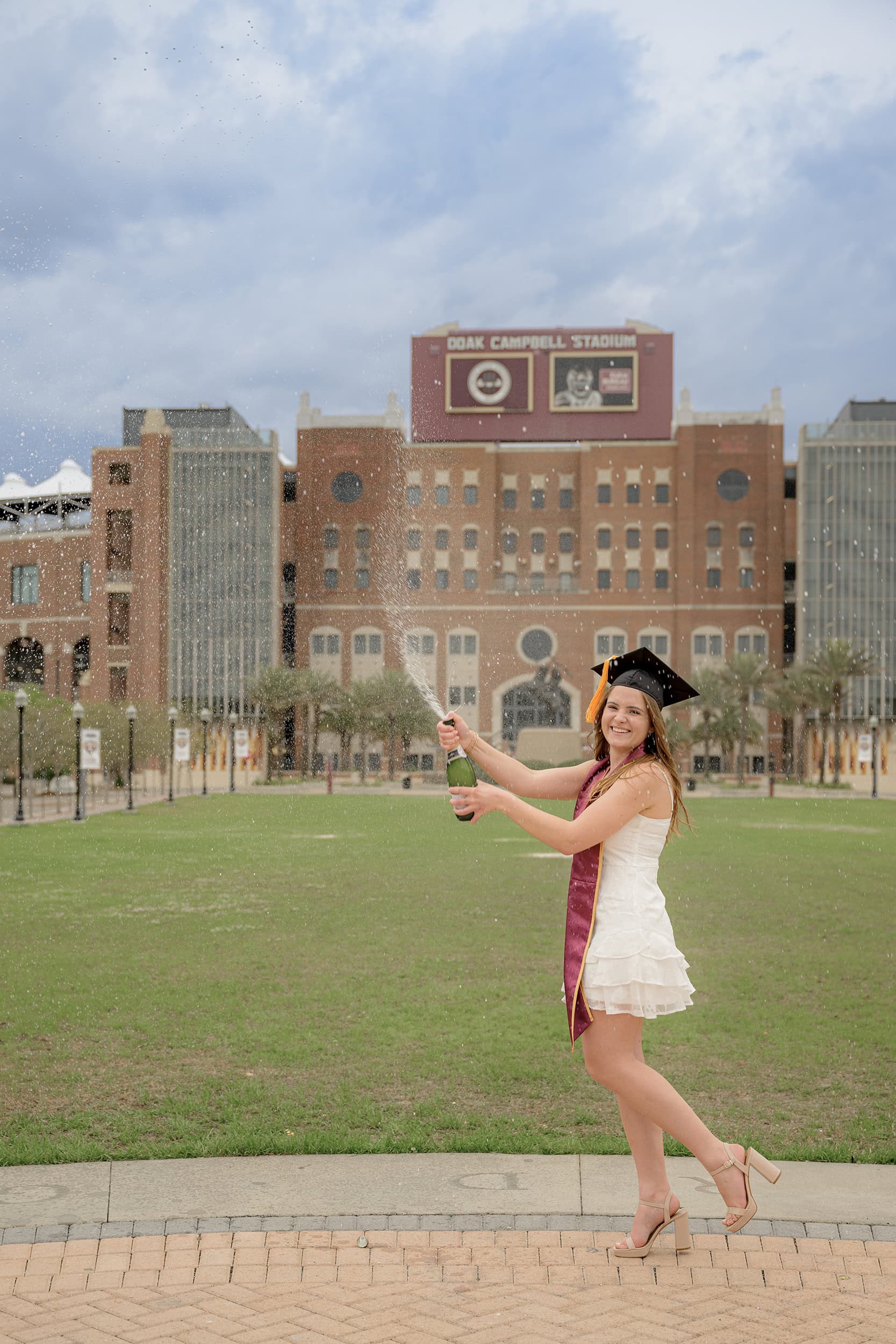 graduate is sprayed with celebratory champagne while standing in front of Florida State University Doak Campbell Stadium wearing her white graduation dress and stole
