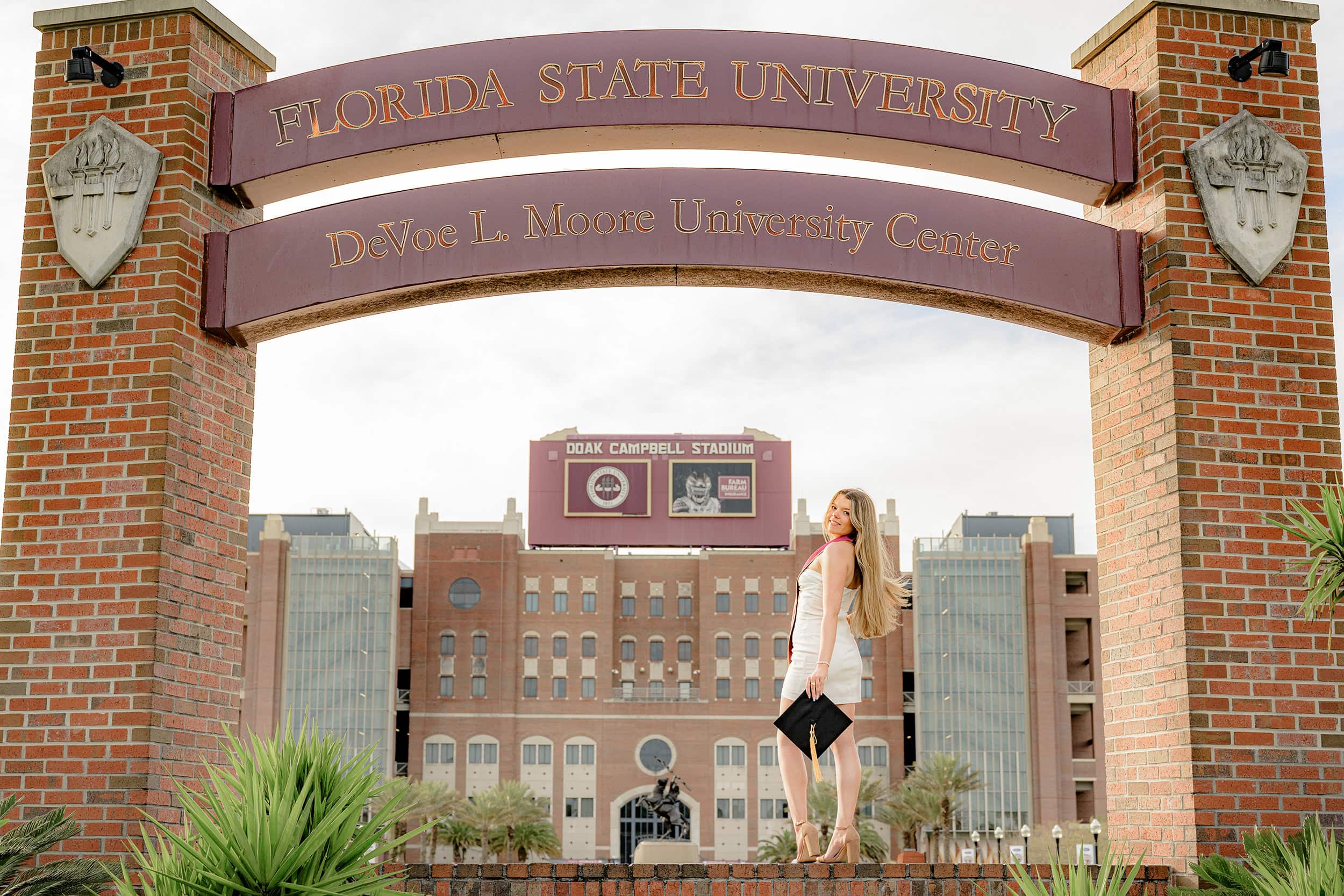 FSU graudate looks back up over her shoulder while standing under the Florida State University sign at Doak Campbell Stadium while holding her graduation cap