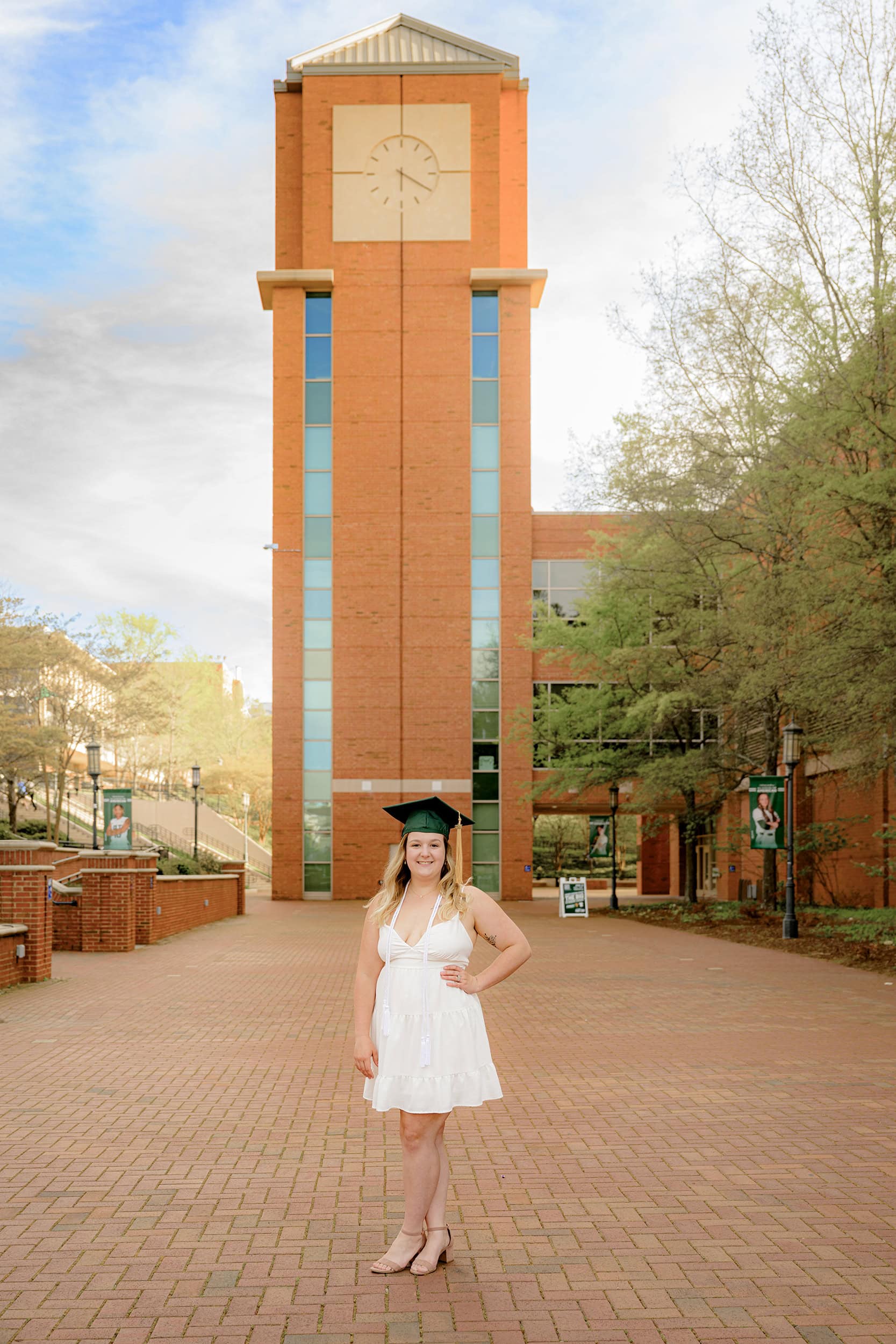 UNCC Senior stands on campus wearing her white graduation dress and cap with tassel in front of clock tower by student union