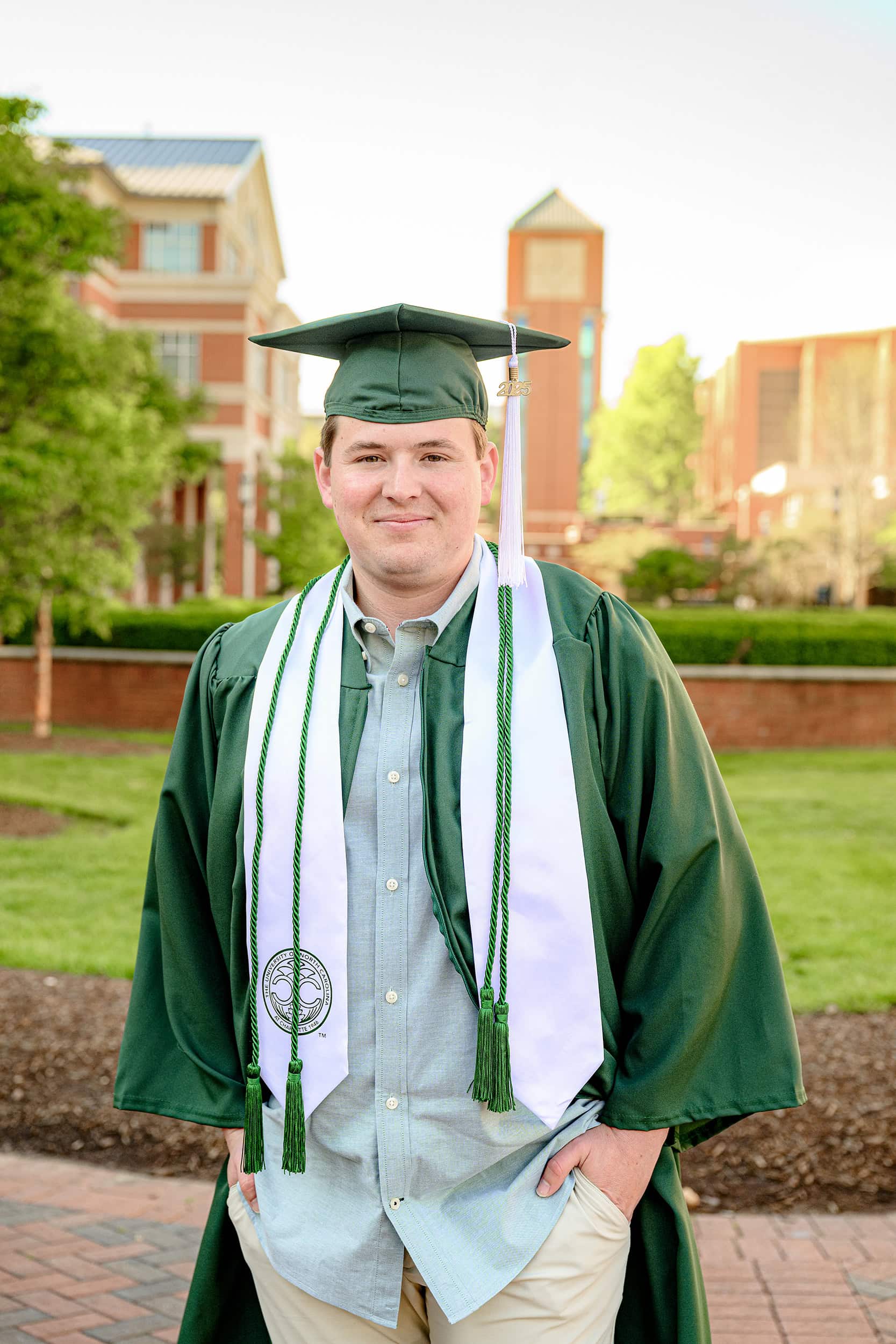 Grad wears University of North Carolina Charlotte green cap and gown with white stole and cords and his graduation cap and tassel standing in front of student union