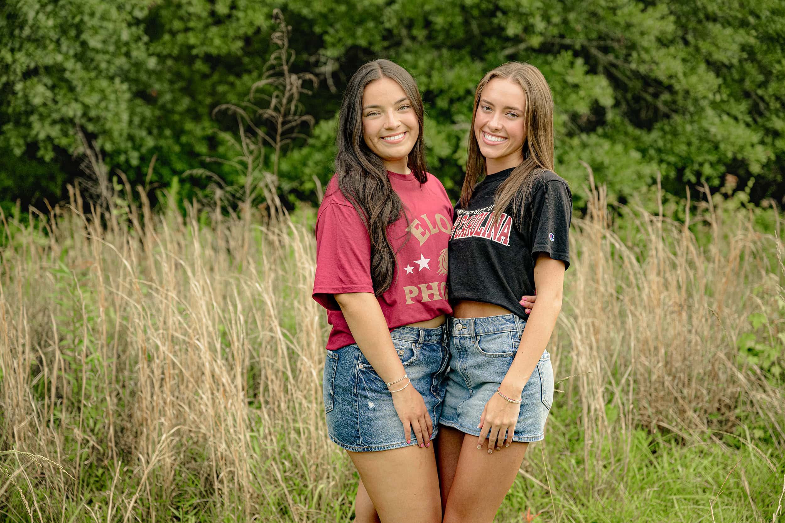 Besties stand together in tall grasses in charlotte area wearing their college tee shirts and jean skirts