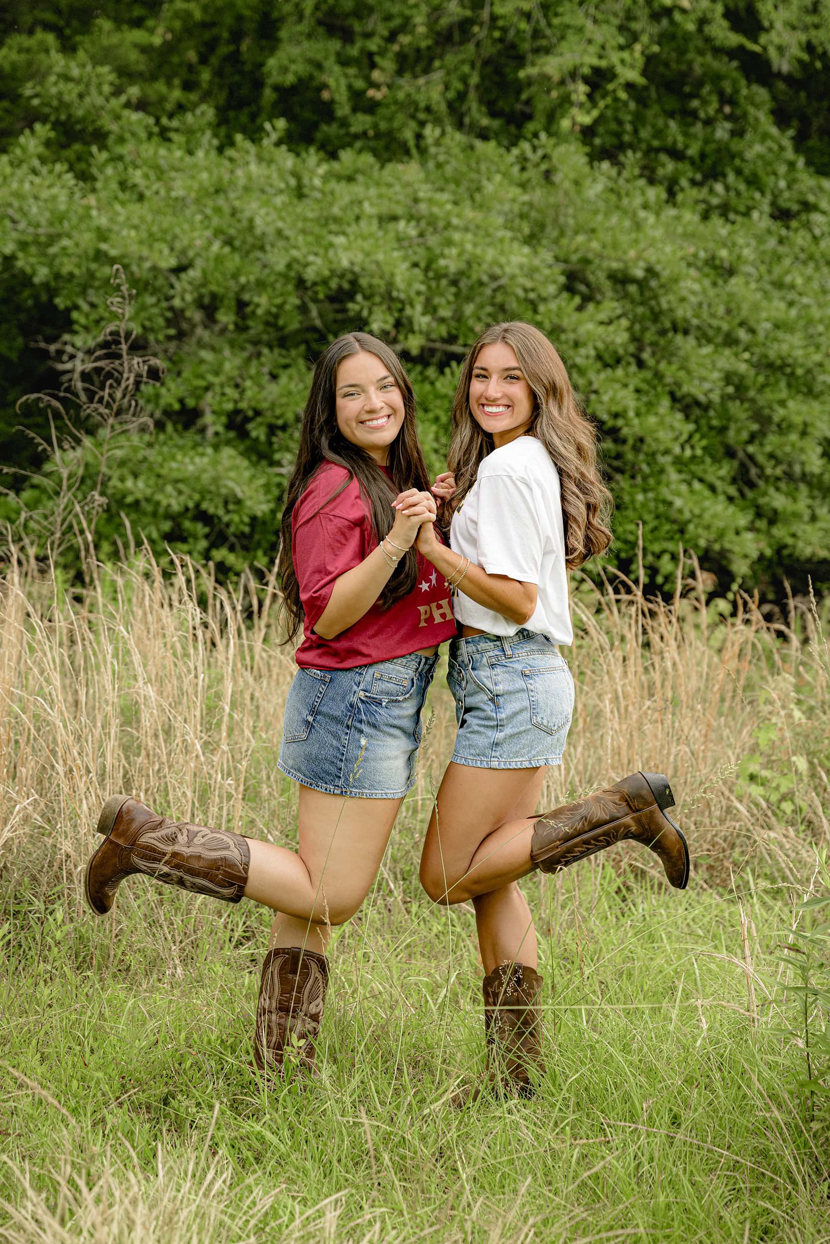 two dark hair besties wearing their college tee shirts, jean skirts, and boots holding hands facing each other in tall grasses