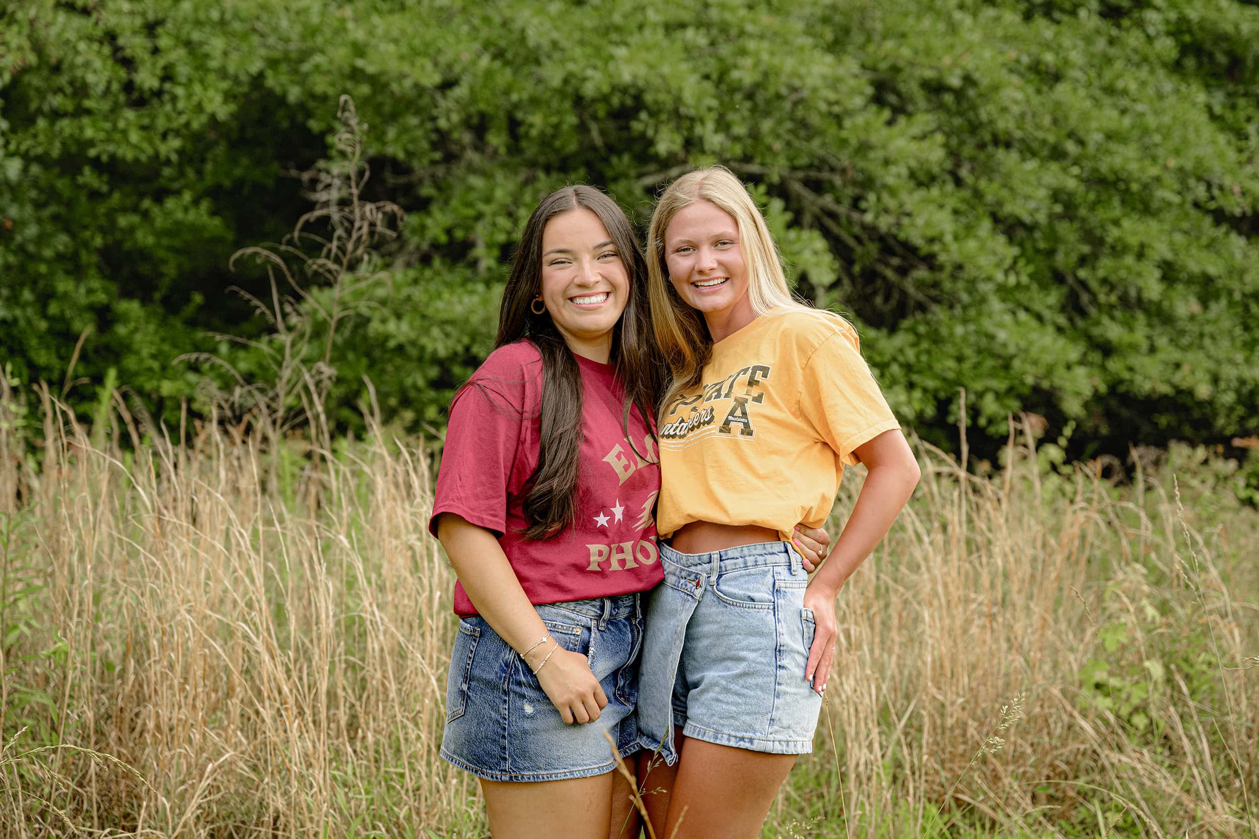 dark hair senior girl wearing her future elon college tee shirt stands next to her bestie in her app state college shirt both wearing jean skirts and standing in tall grasses