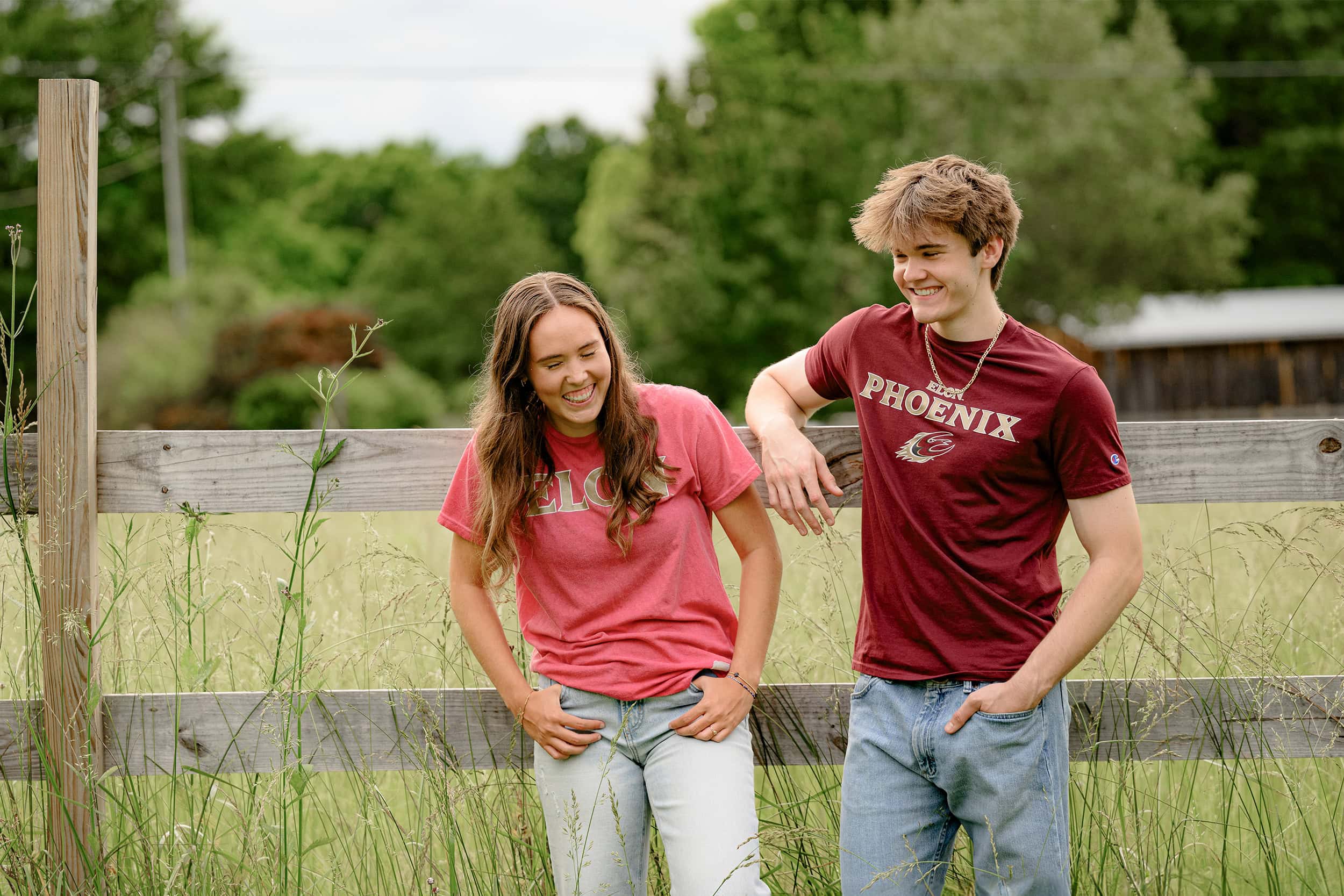 twin sister wearing pink elon univeristy shirt laughs as she stands next to her twin brother wearing a maroon elon phoenix shirt while leaning against a fence in a charlotte field