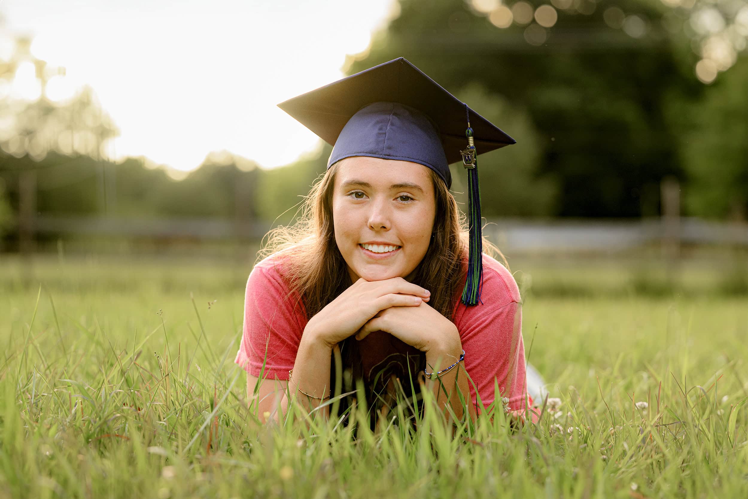 twin girl lays in field of tall grasses wearing her weddington high school cap and tassel during her twin bestie session