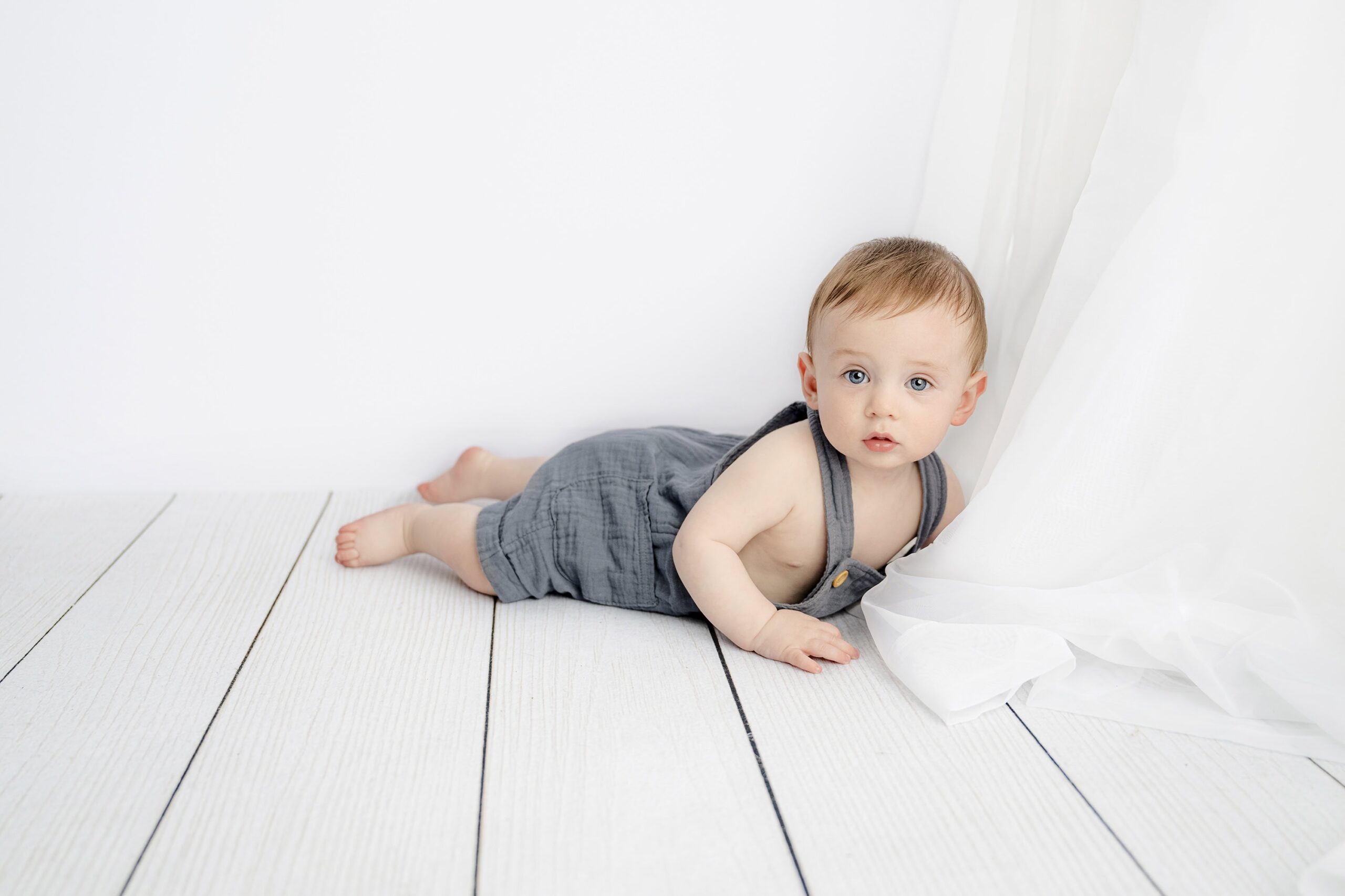Eight month old boy laying on his belly wearing light blue overalls