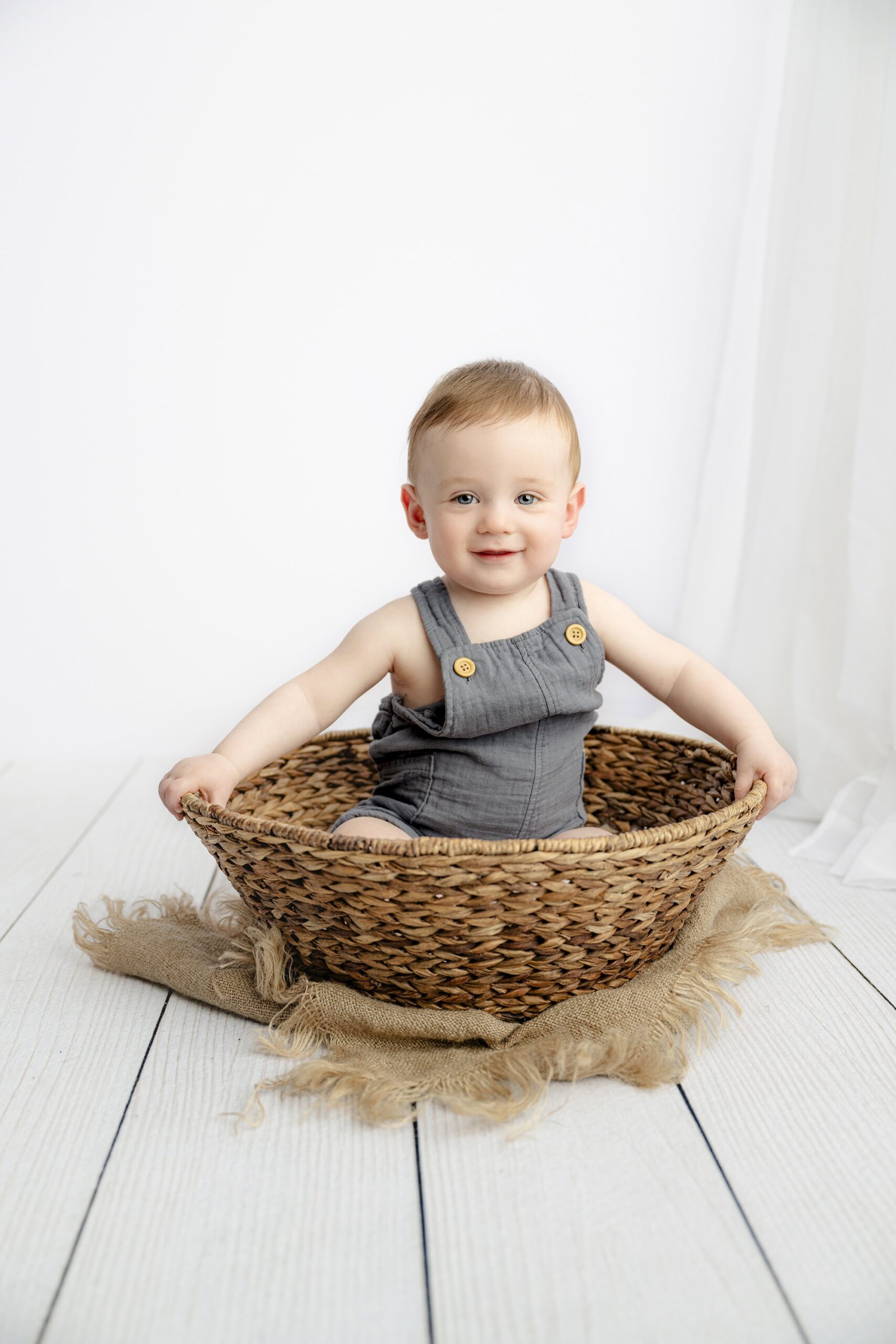 little 8 month old boy sitting in a brown basket wearing blue overalls