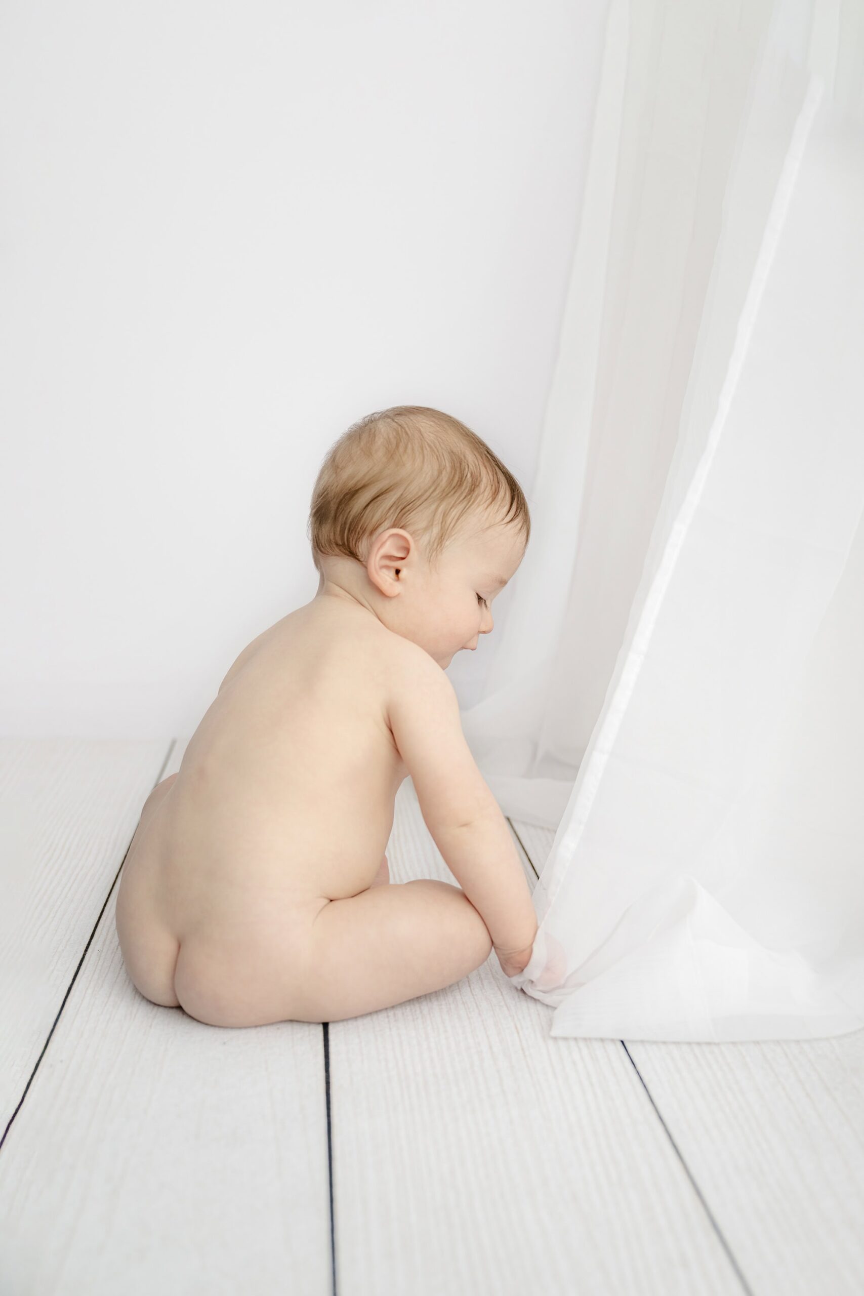 proof babies play with anything as this little one sans diaper and playing with white studio curtain in a waxhaw milestone studio