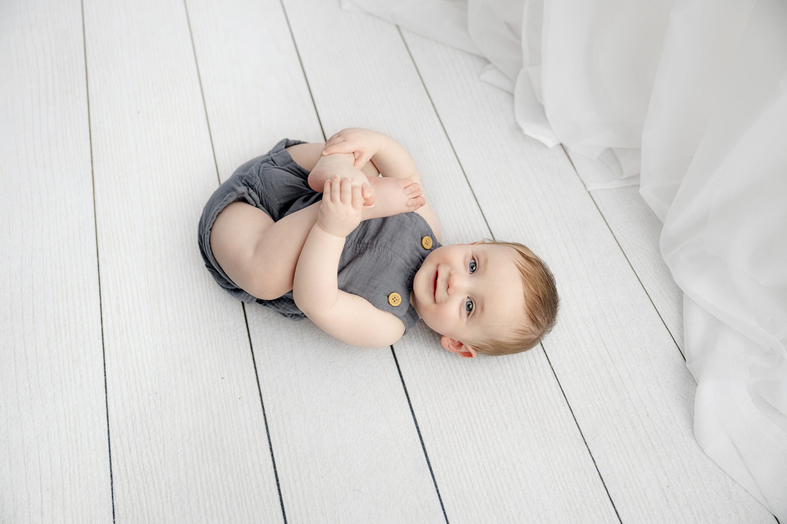 rolling over on his back playing with his feet wearing blue overalls in a white studio set