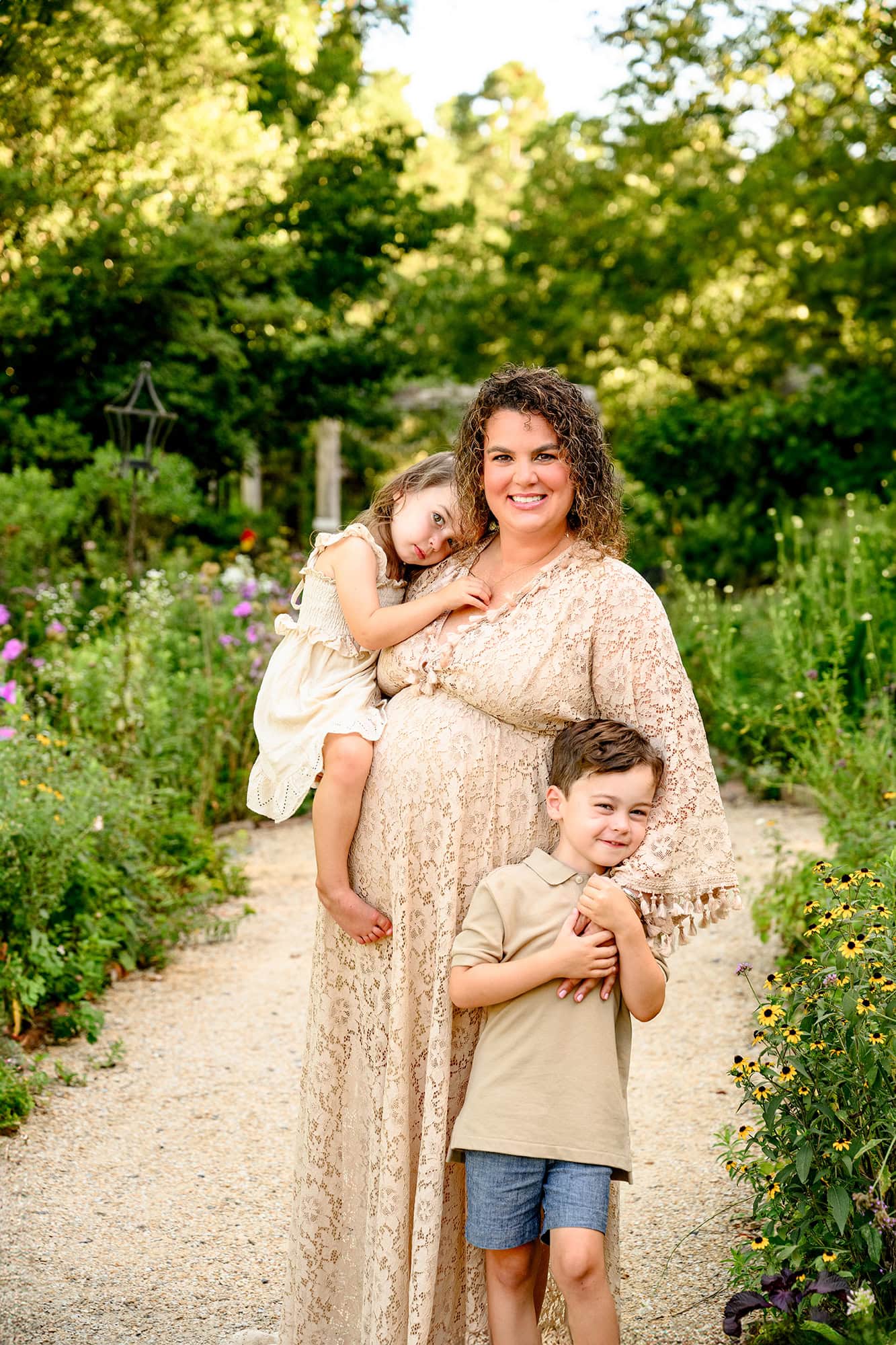 mom to be holds daughter with her arm around her son standing while pregnant in a charlotte area flower garden with little sister holding 3d ultrasound