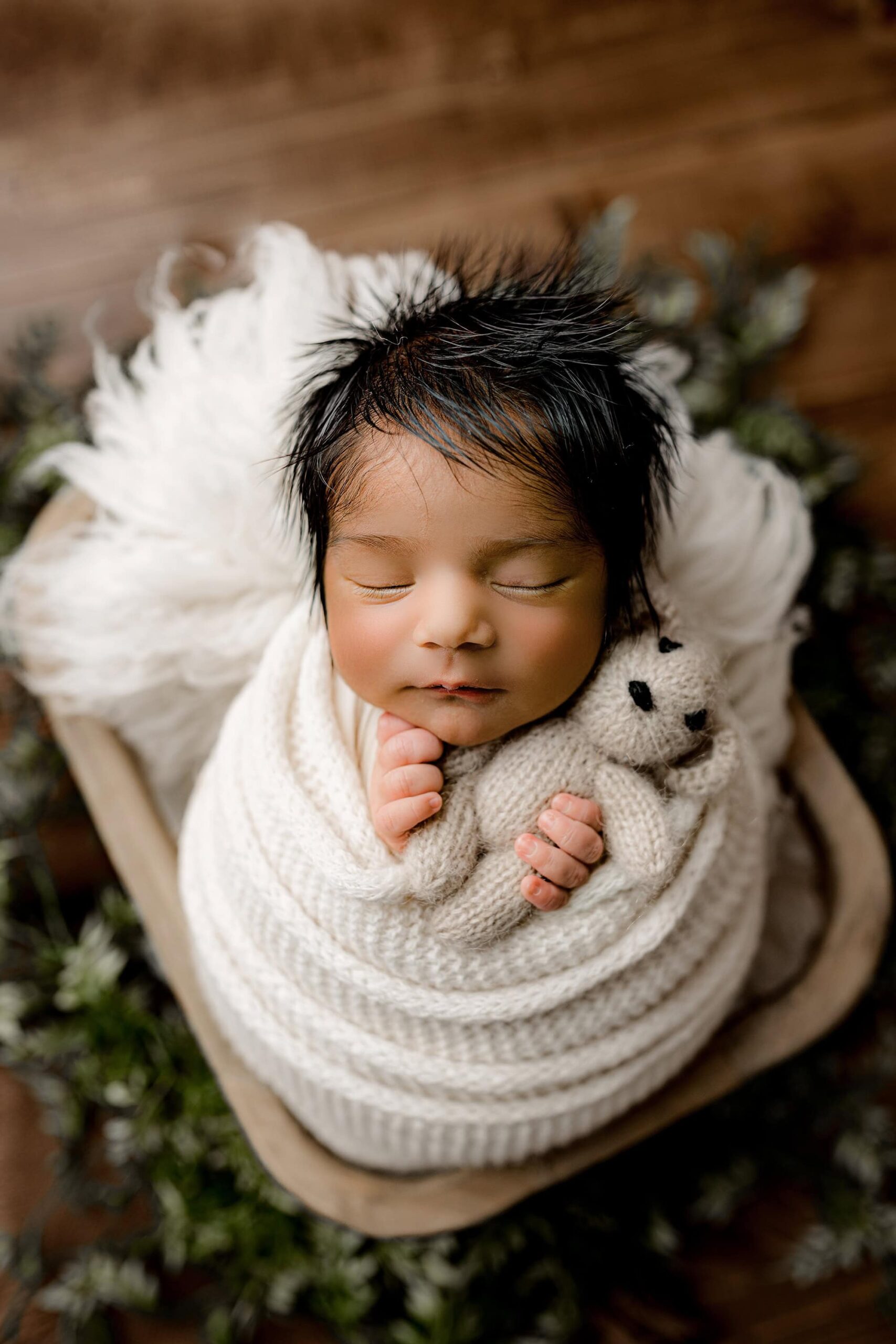 newborn baby boy is wrapped in a cream wrap holding stuffy sitting in a crate