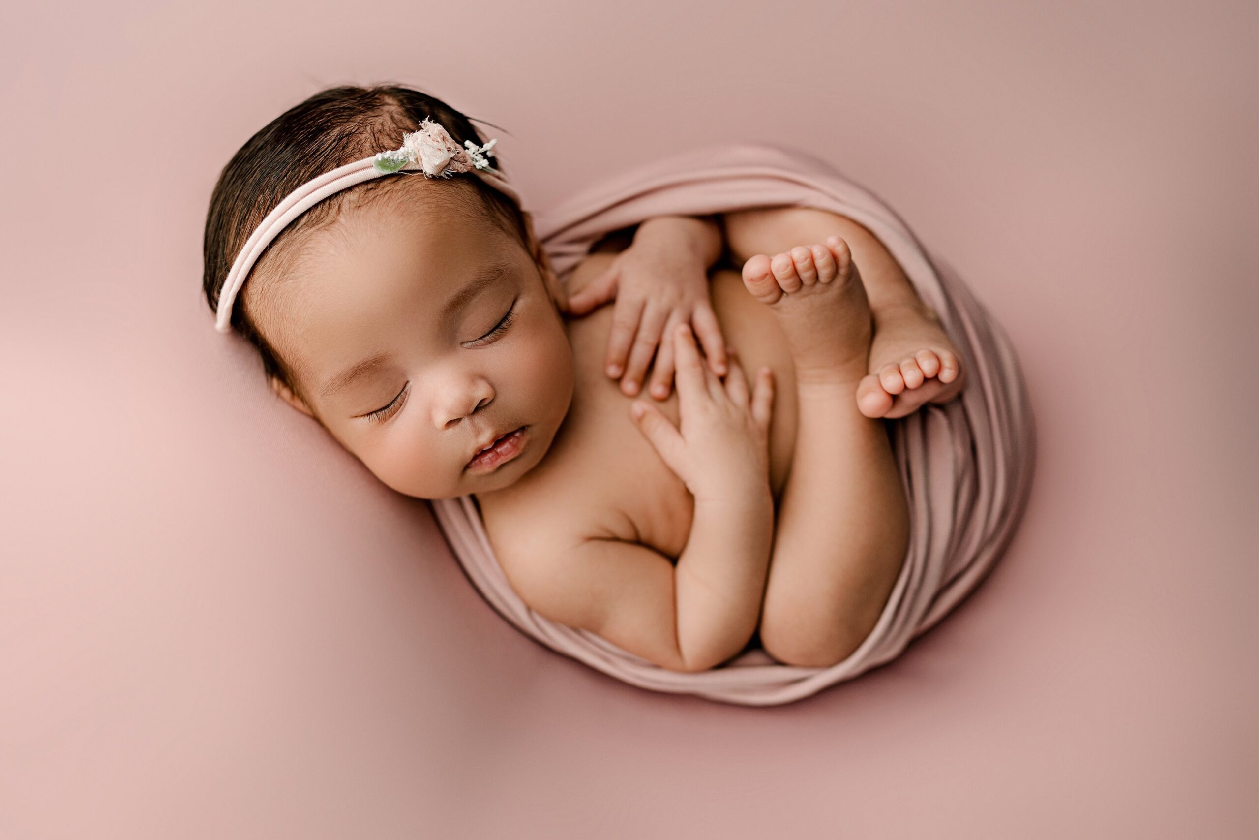 newborn baby girl lays on mauve background wrapped in complimentary colored wrap and matching bow