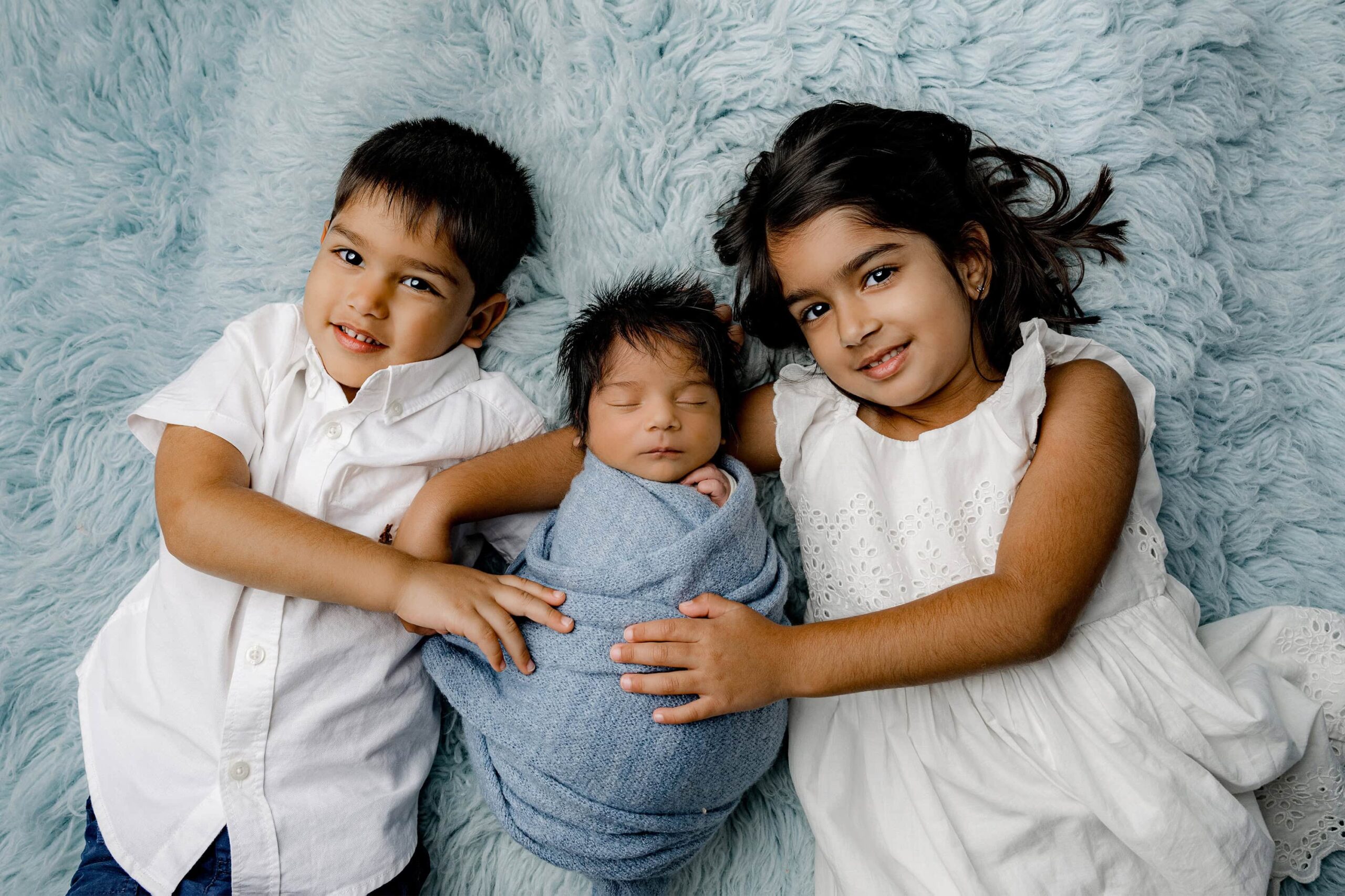 two dark hair older siblings dressed in white lay on their backs holding their newborn brother wrapped in blue.