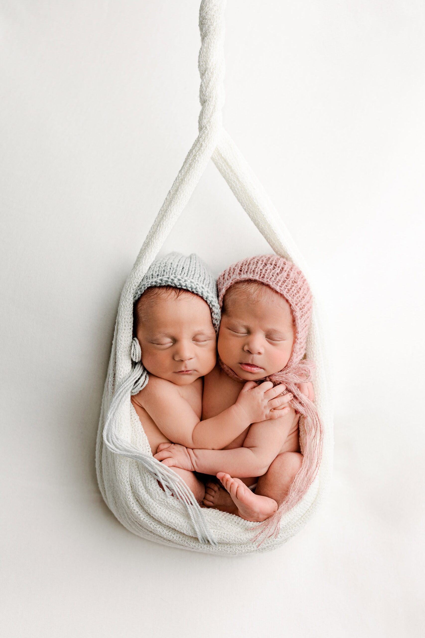 twin newborn boy girl siblings cuddled together wearing a blue and pink bonnet wrapped in a white wrap