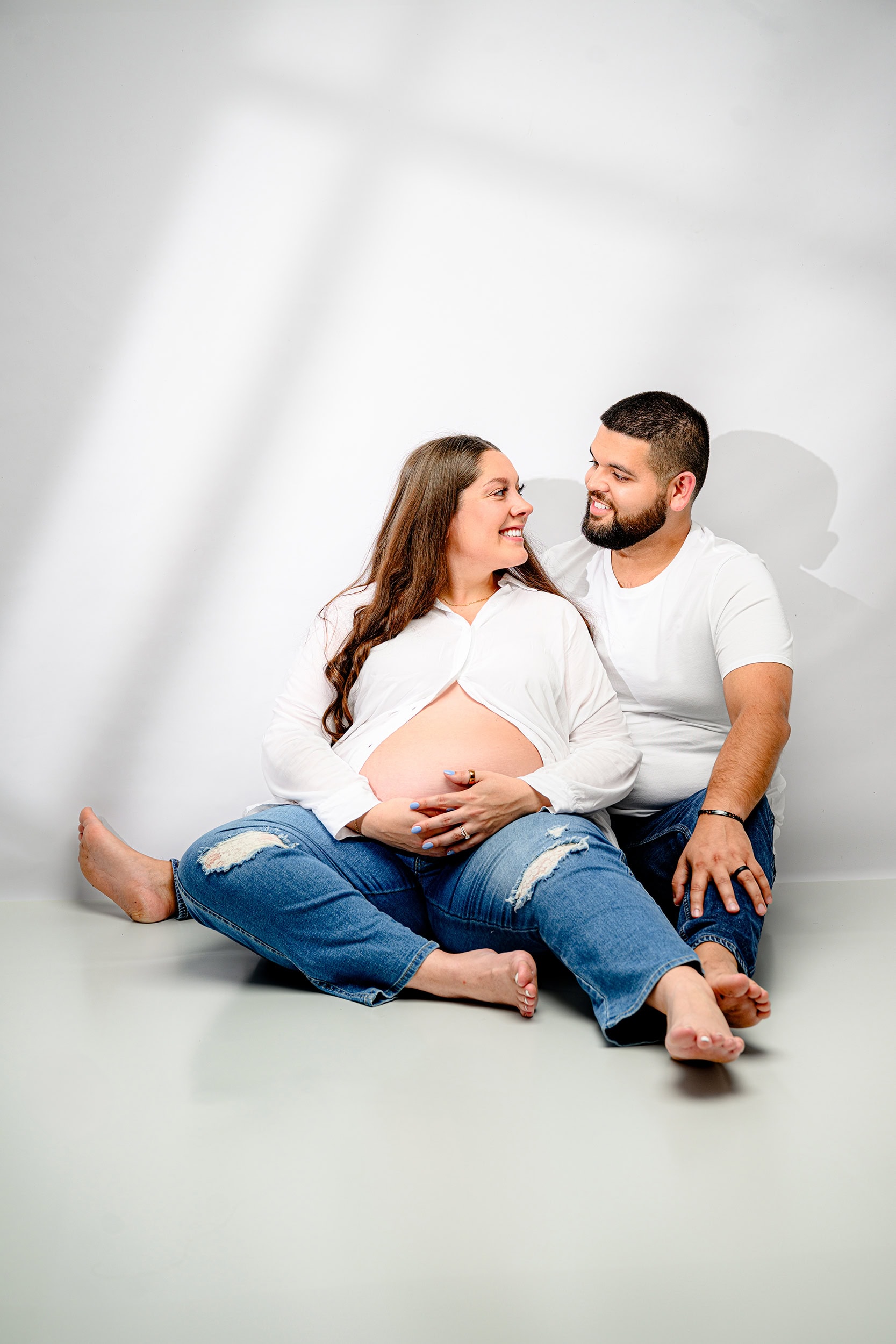 expectant couple dressed in white shirts and jeans sits in a waxhaw studio looking at one another smiling with mom holding her belly
