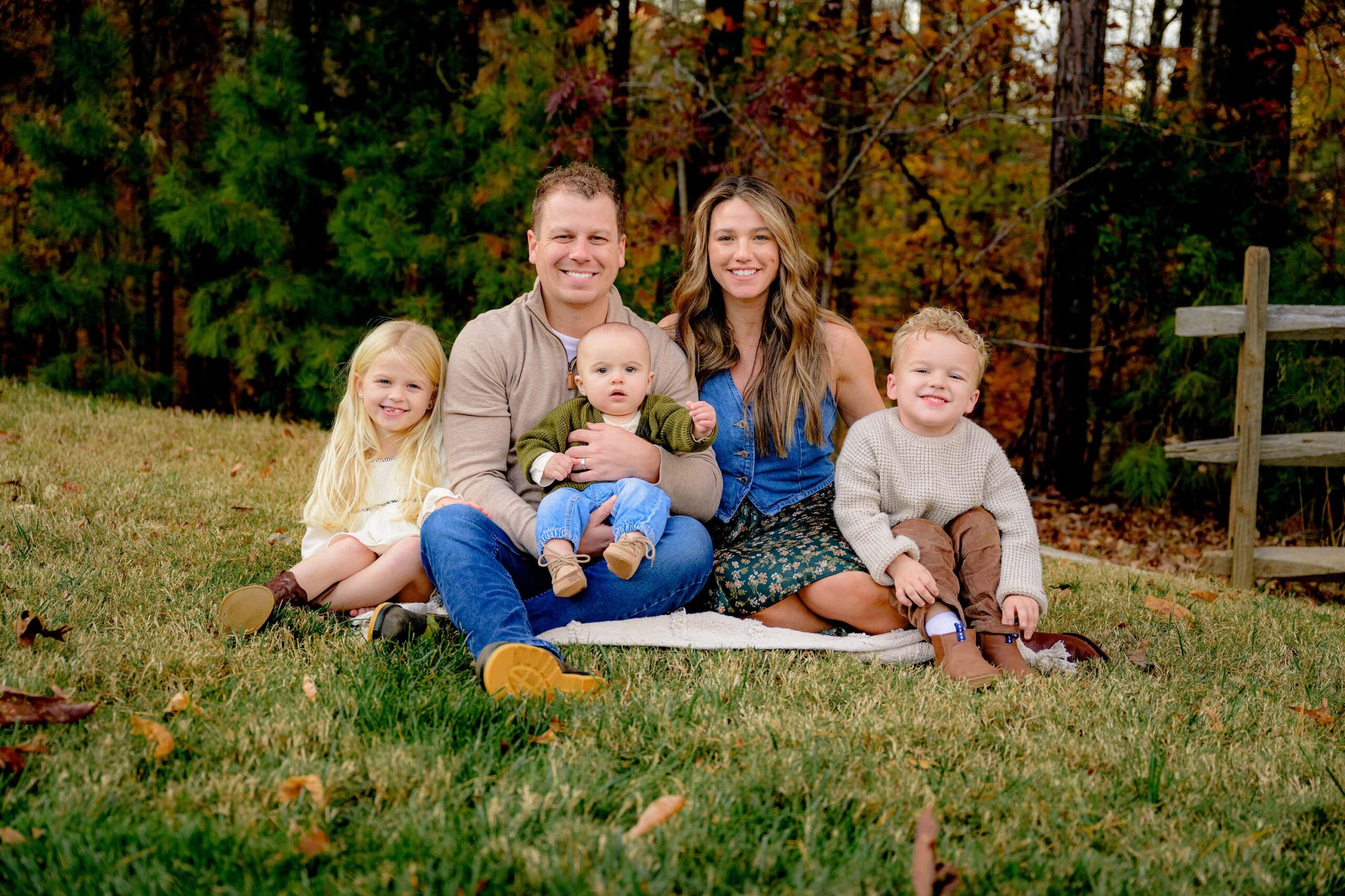 Family of 5 - two parents and 3 kids sit together in a grassy fall location with fall trees behind them