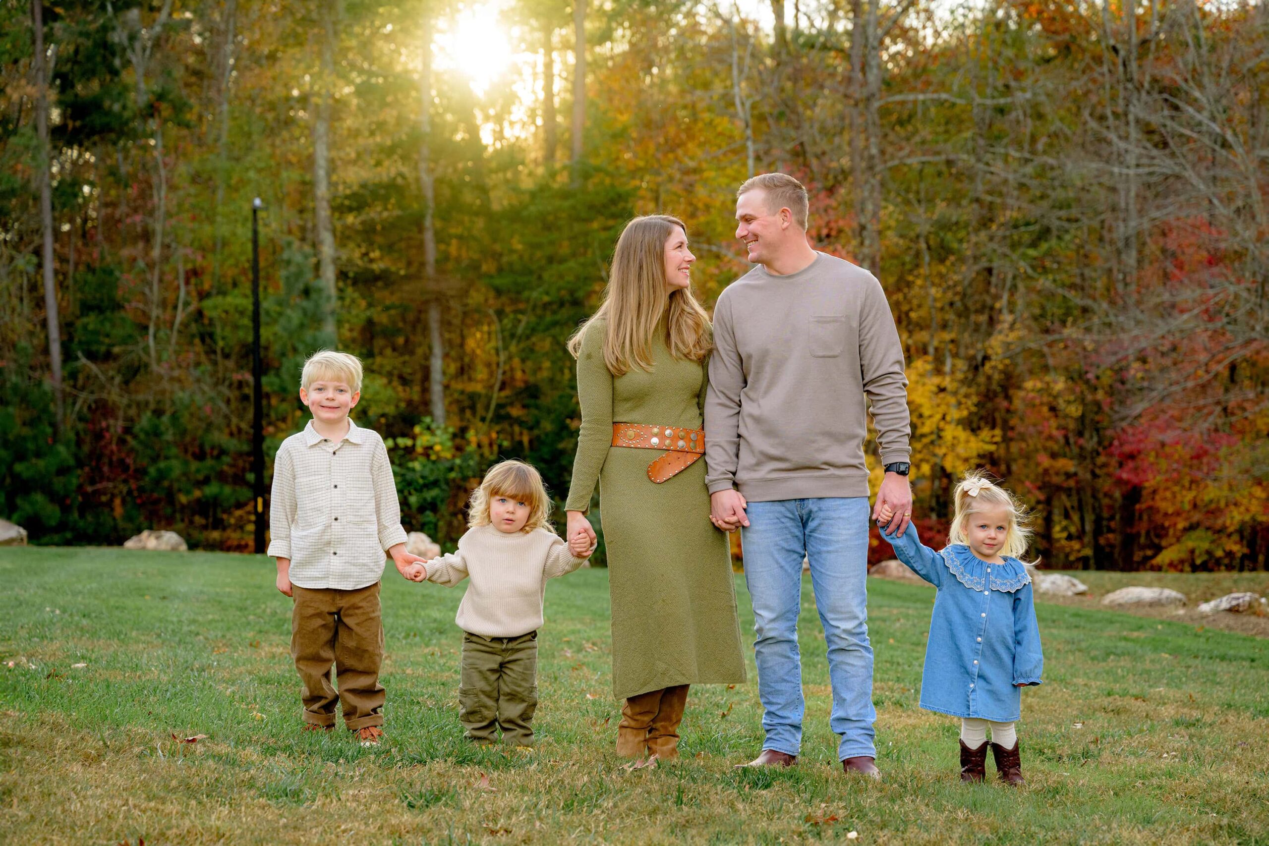 two parents look at one another while holding hands with their children with the autumn sun behind them after attending a fall festival