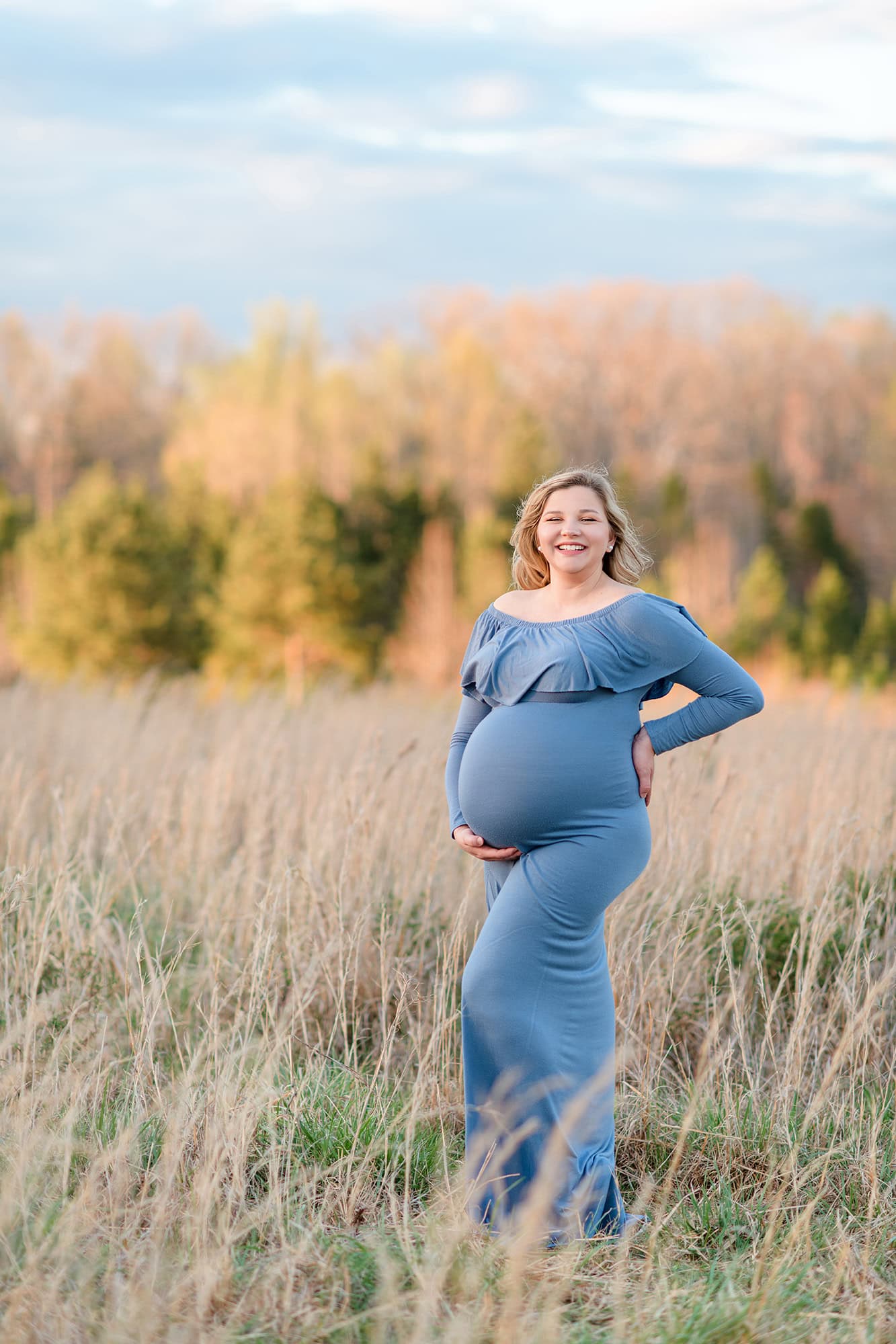 expectant mom stands in tall grasses laughing while dressed in a blue maternity dress surrounded by a charlotte fall scene