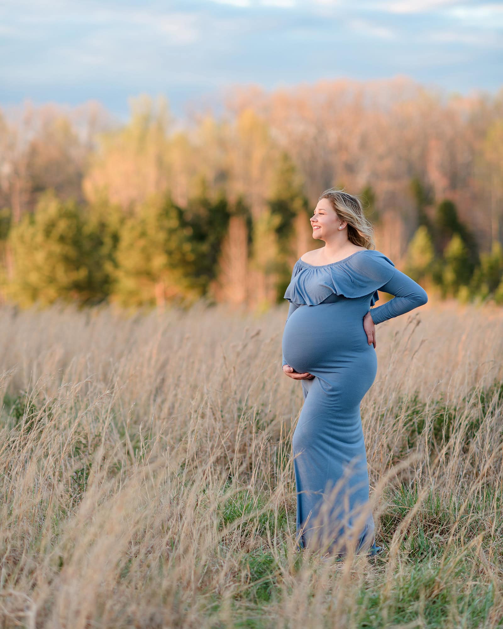 Pregnant mom stands in tall grass field in fall wearing a blue dress surrounded by charlotte fall colors