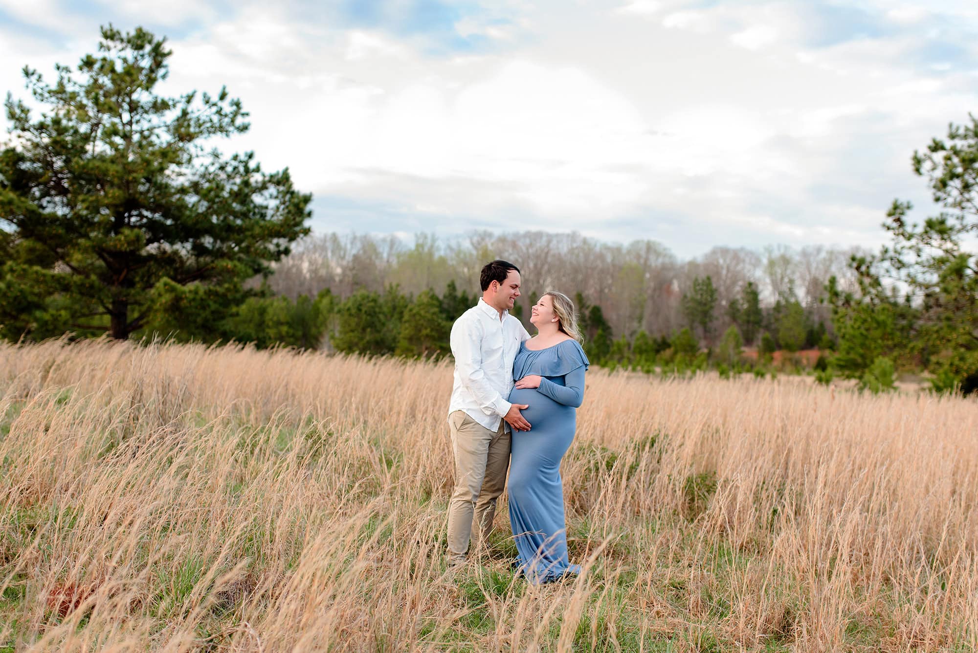 Charlotte expectant couple poses together in a fall field with mom wearing blue dress and dad in a white shirt