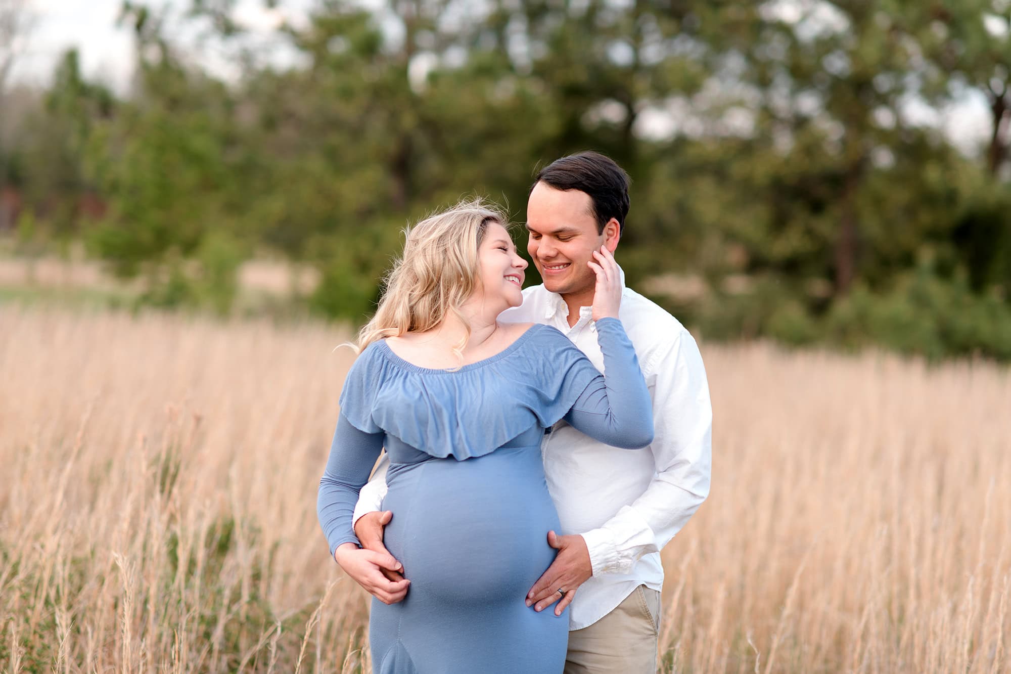 mom touches dad's face as he hugs her expectant belly while laughing together during maternity photos