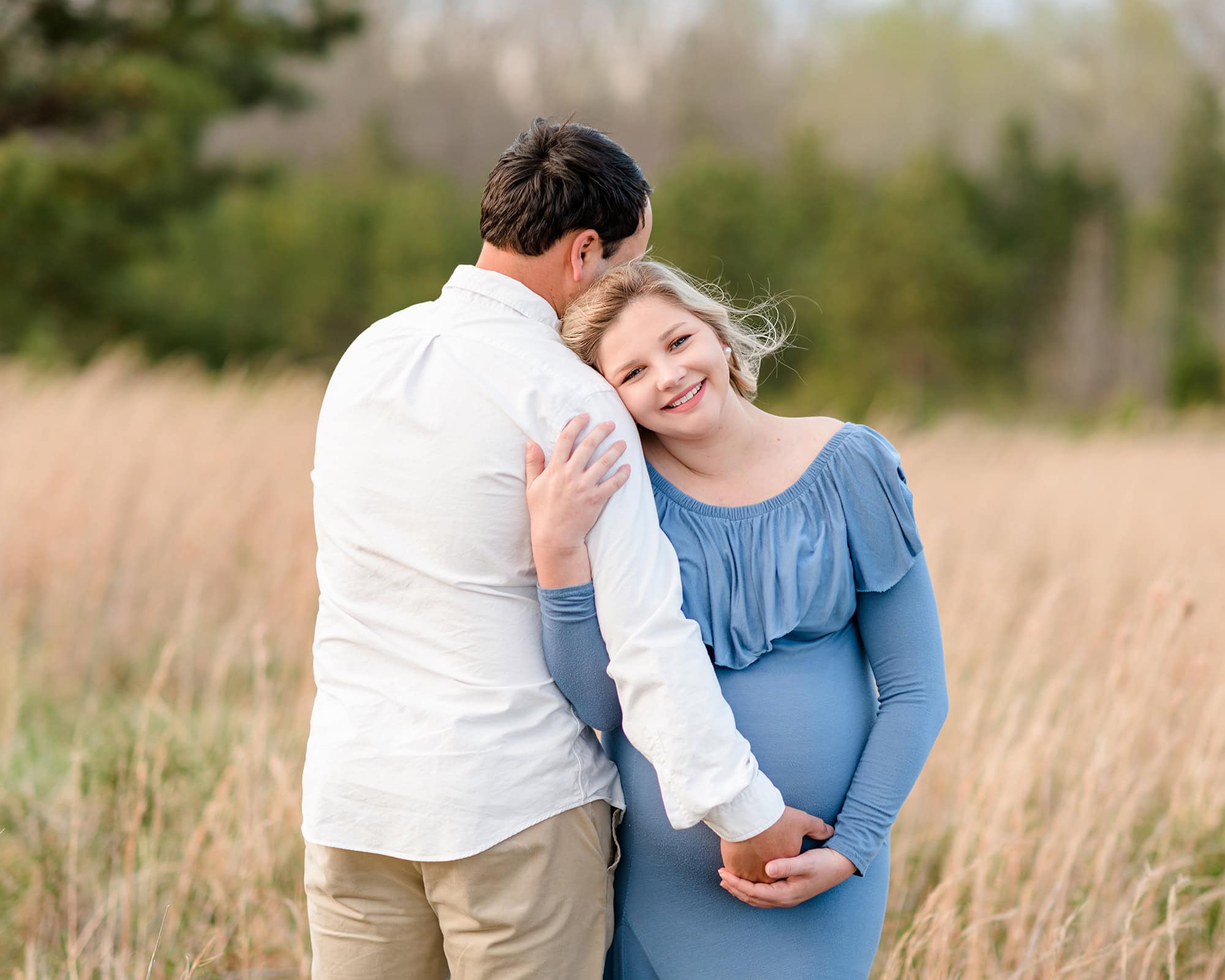 pregnant mother leans against her husband in a fall field wearing a blue dress