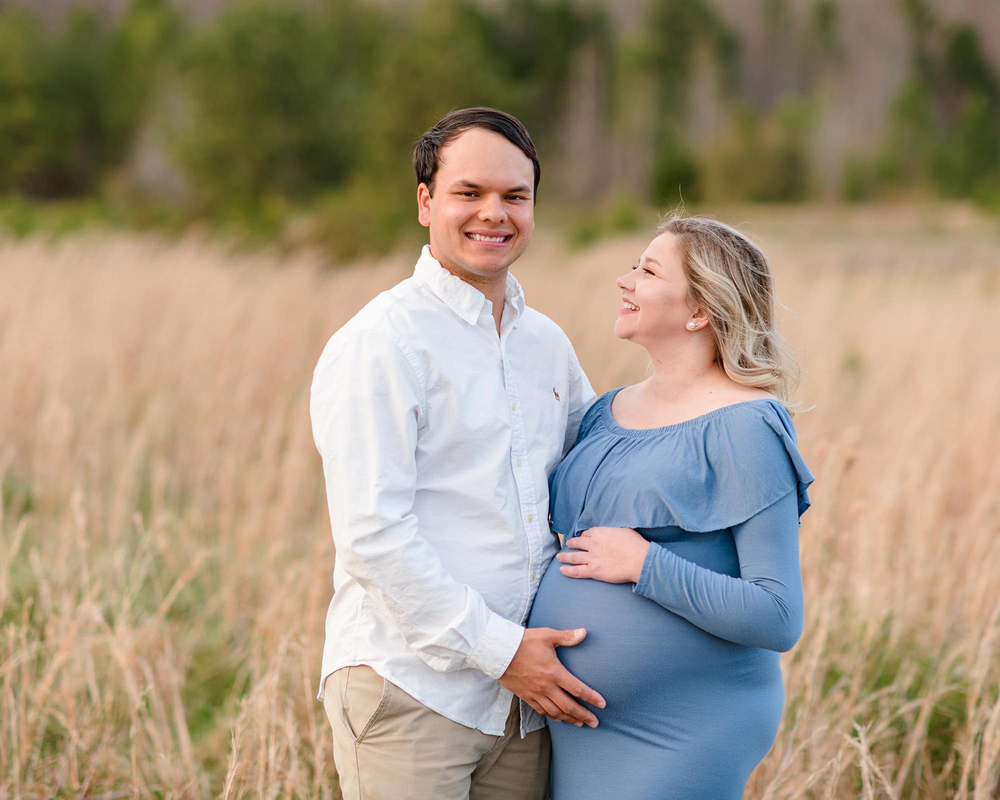 pregnant mom looks at her husband while standing in a fall field and both with hands on her belly