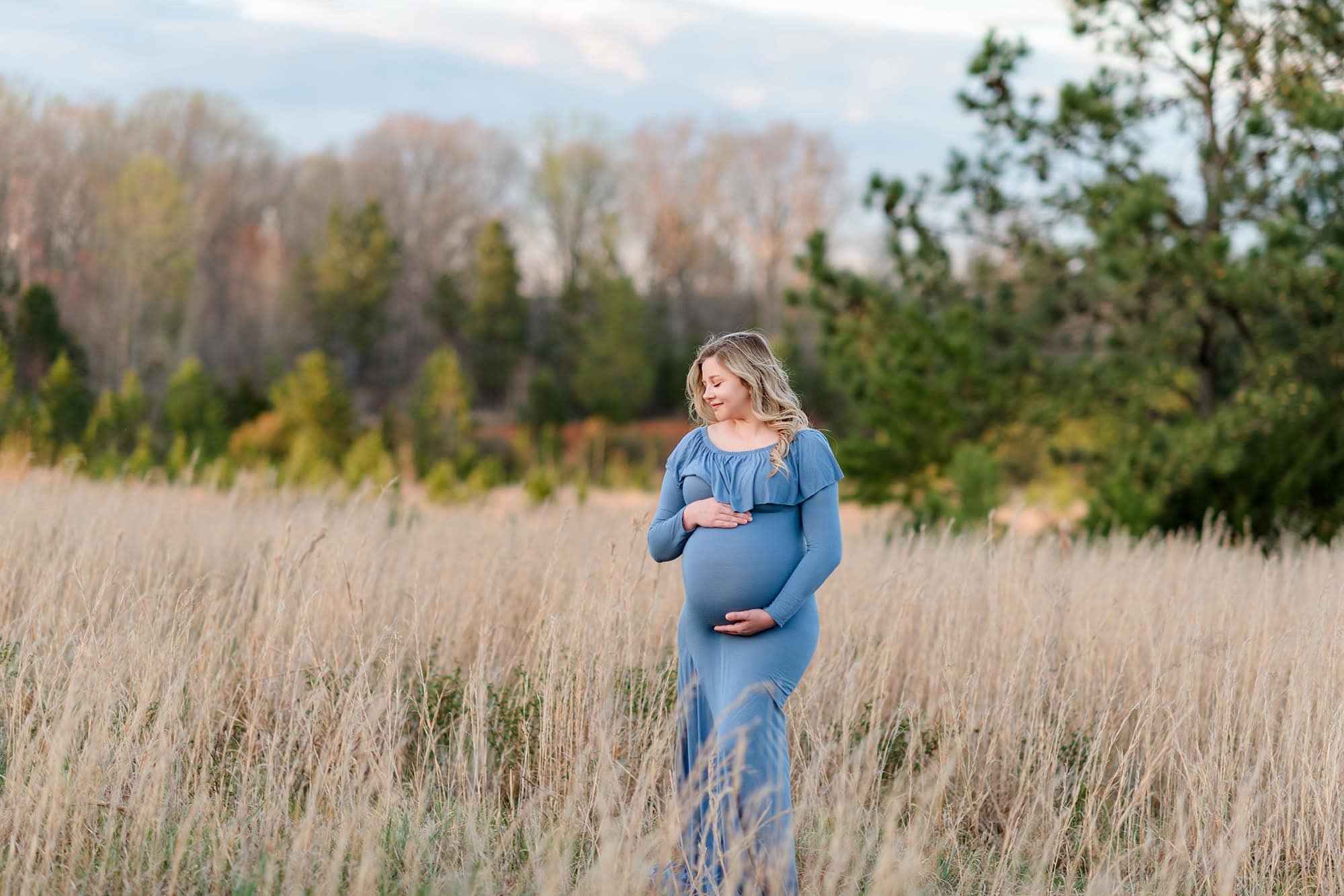 expectant mom looks downward while standing in a fall field of tall grasses wearing a blue dress