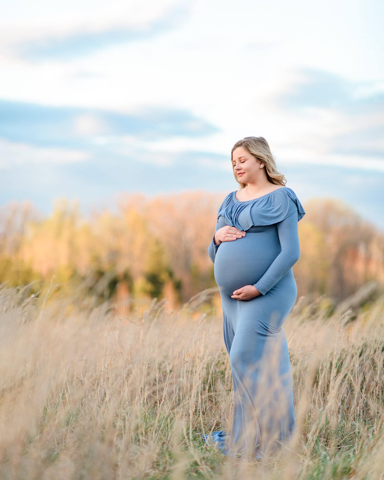 fall tones in the background of this charlotte area location with a pregnant mom standing in tall grasses cradling her belly wearing a blue dress