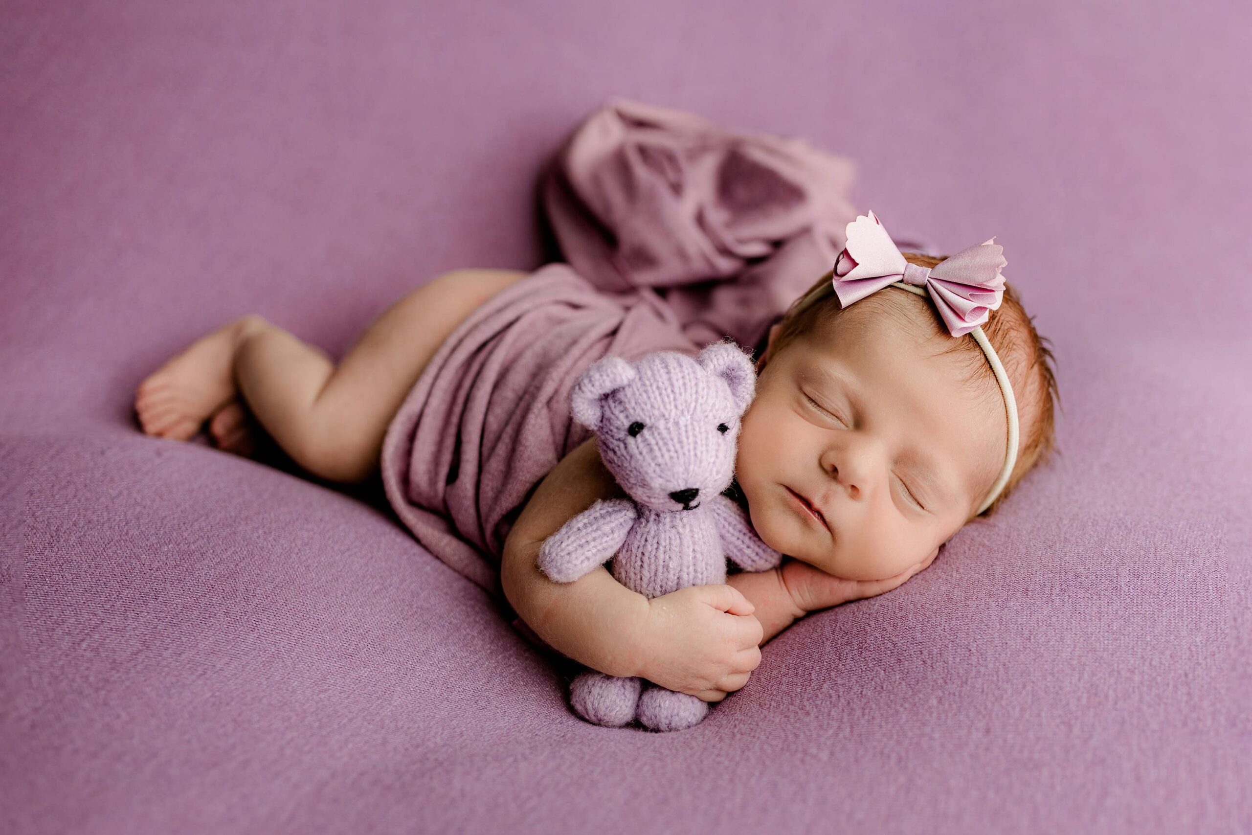 Charlotte infant girl lays on her side with one hand tucked under her cheek and one arm holding a purple teddy bear on a purple background perfect for her woodland nursery theme