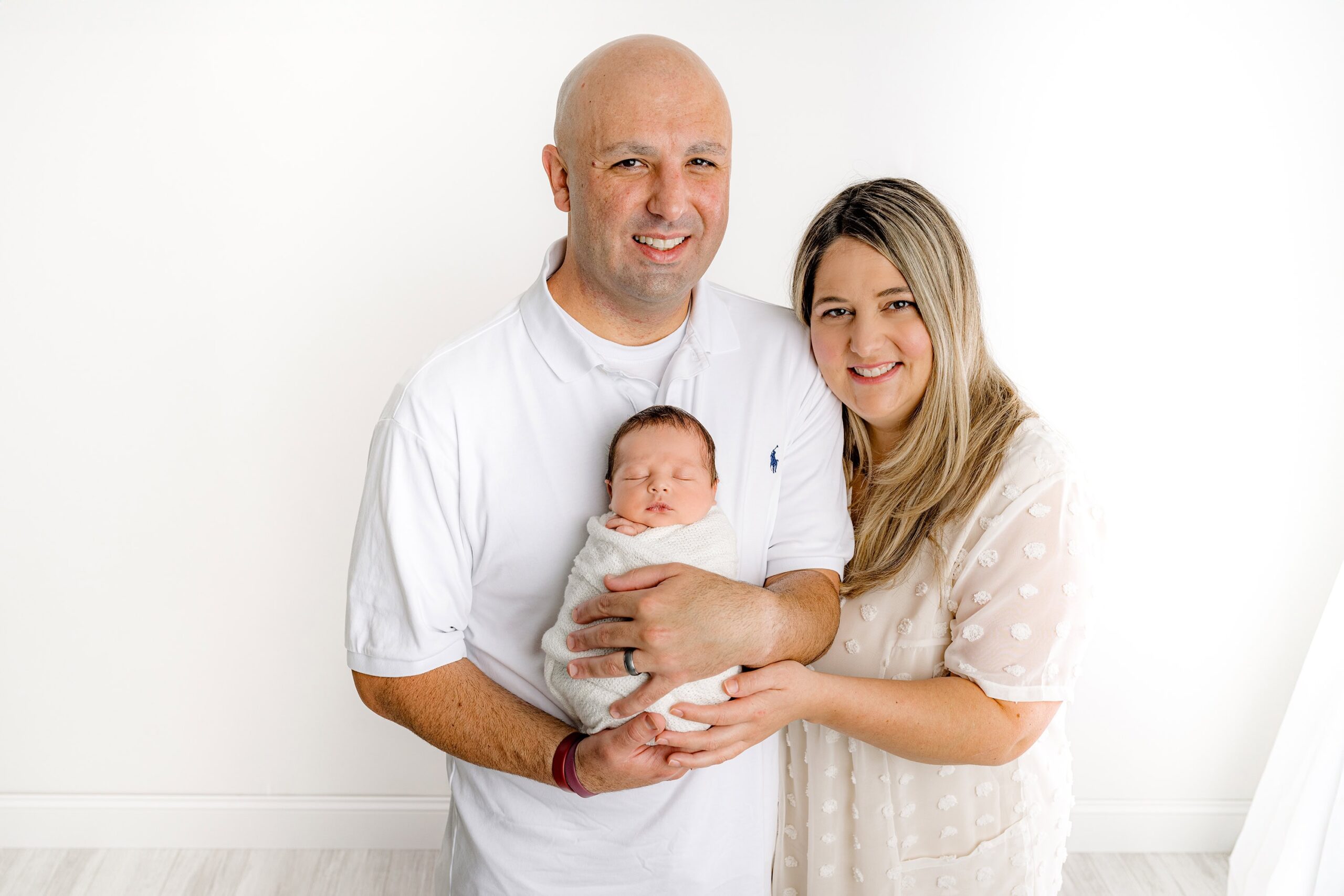 Father dressed in white holds his newborn son in this waxhaw studio portrait session with mom standing beside the duo