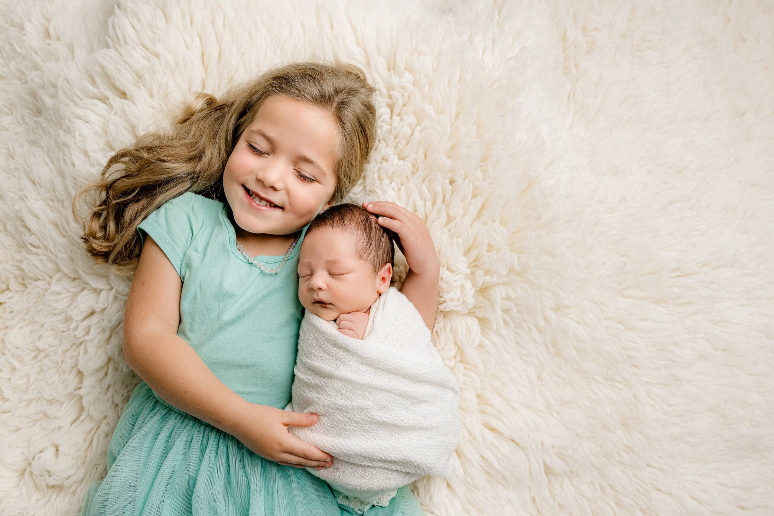 little girl dressed in pool blue colored dress lays on her back cradling her baby brother wrapped in white