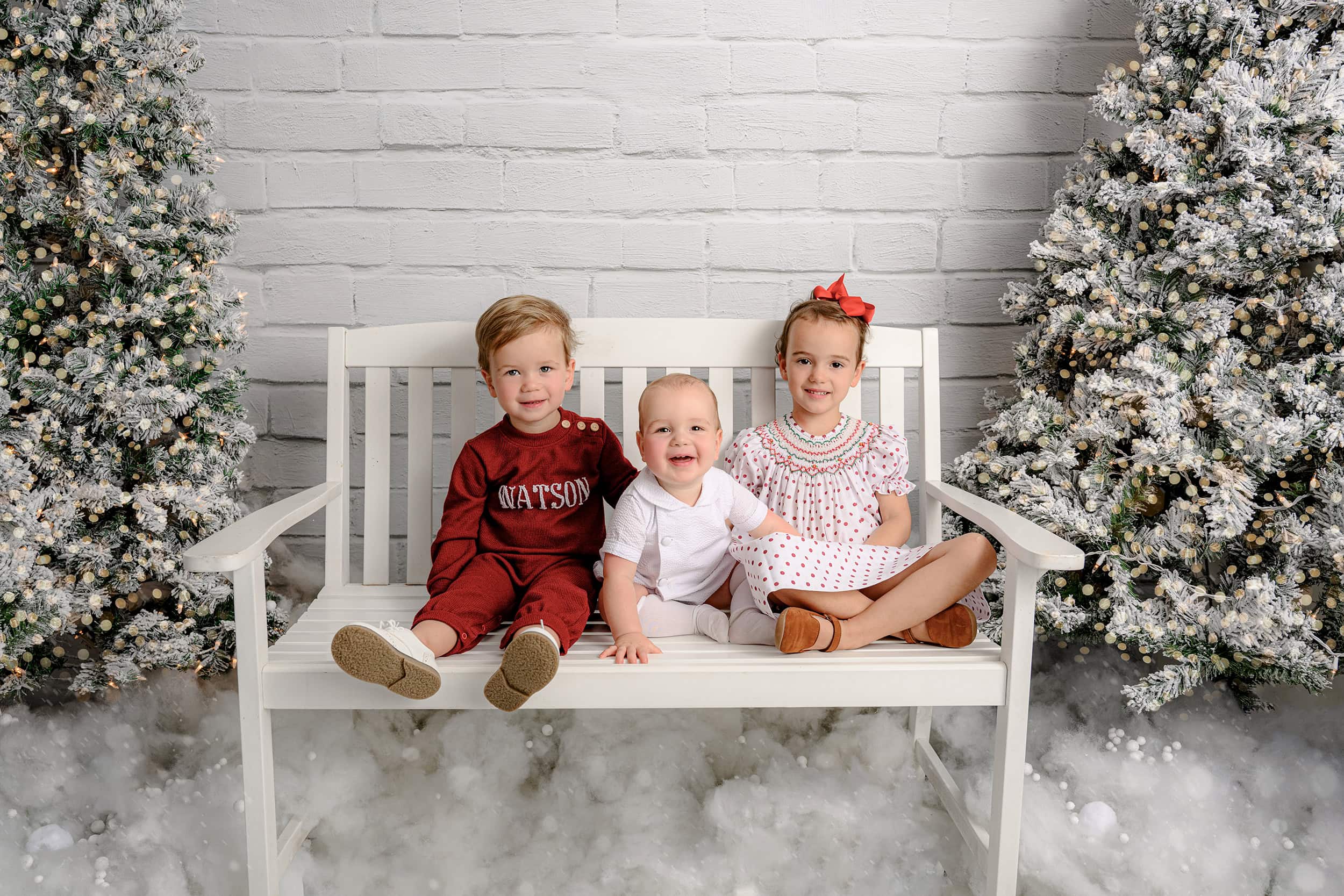 three siblings from the charlotte area all dressed in chrismtas colors surrounded by white holiday trees 