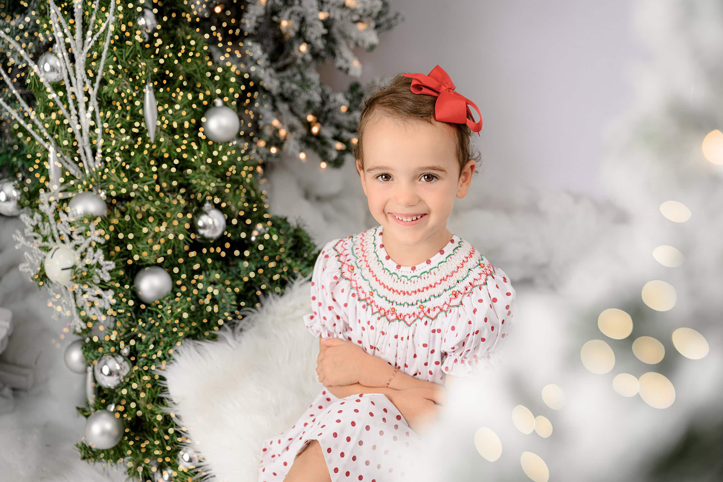 little waxhaw girl in holiday dot dress and wearing a bright red bow sits in a garland covered studio set up 