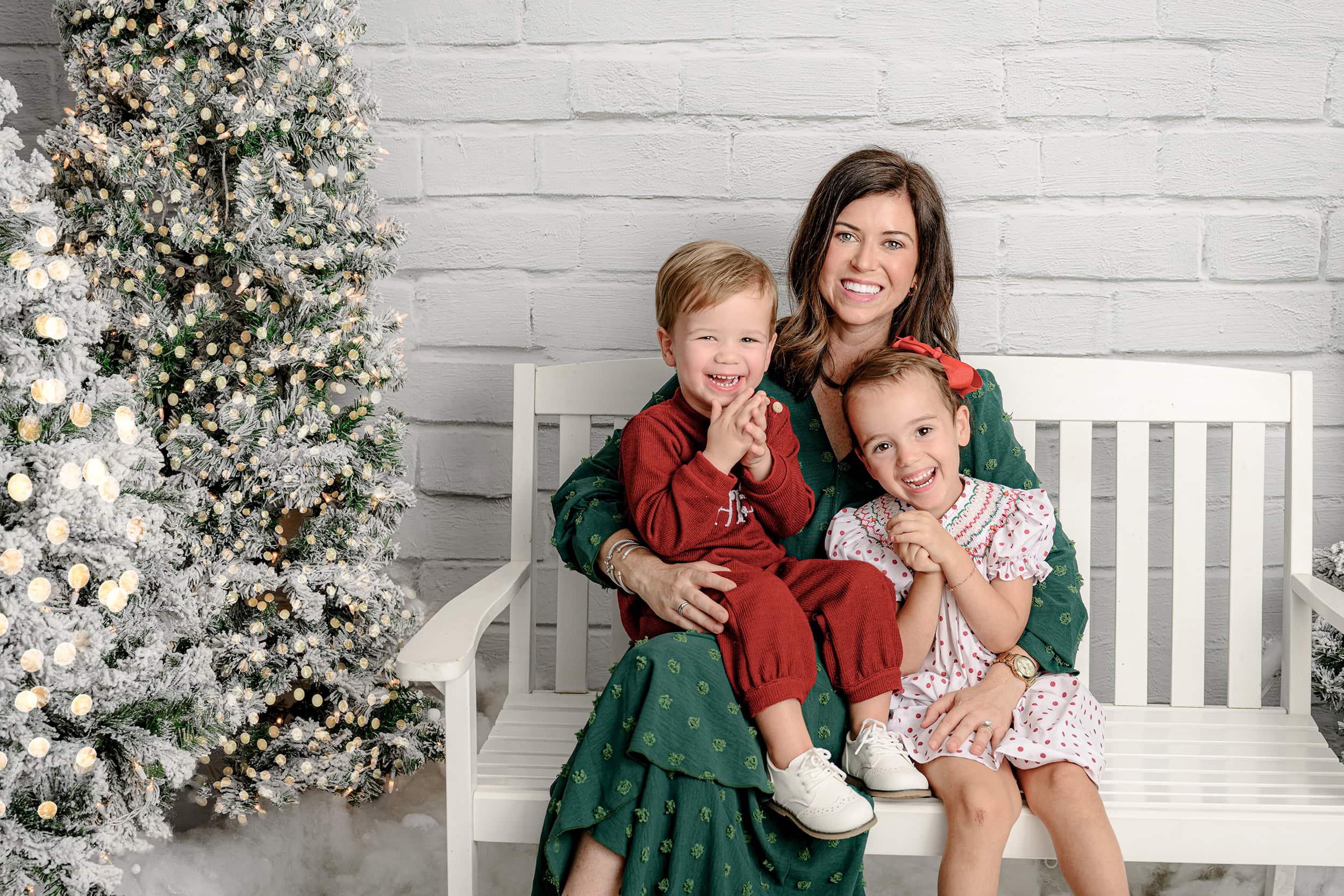 two children sit in their mother lap laughing while dressed in holiday colors surrounded by white christmas trees
