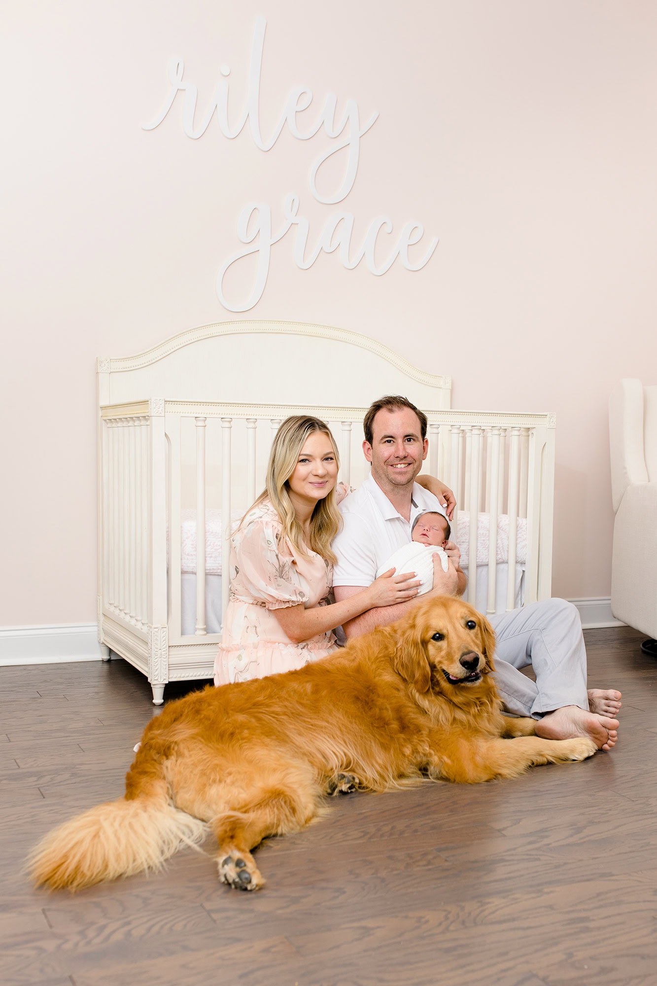 parents sit holding their newborn girl by her crib with their dog beside them