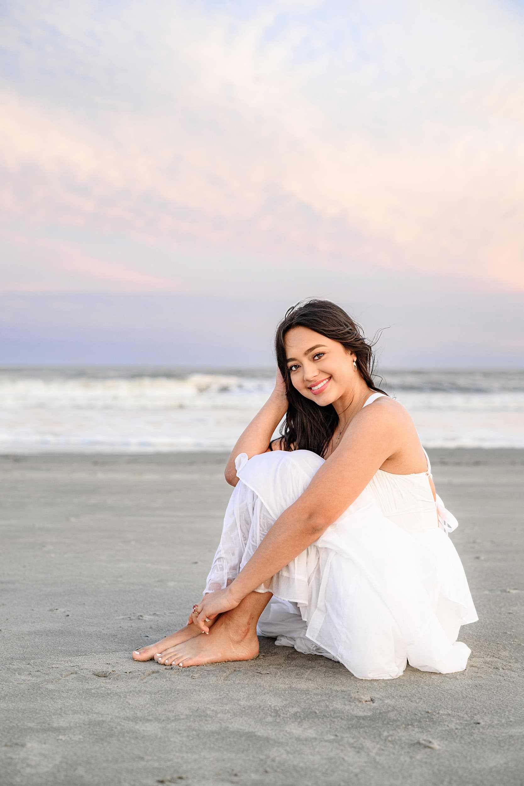 dark hair charlotte graduate sits on the beach in white dress from shop iris and poppy in downtown waxhaw, nc