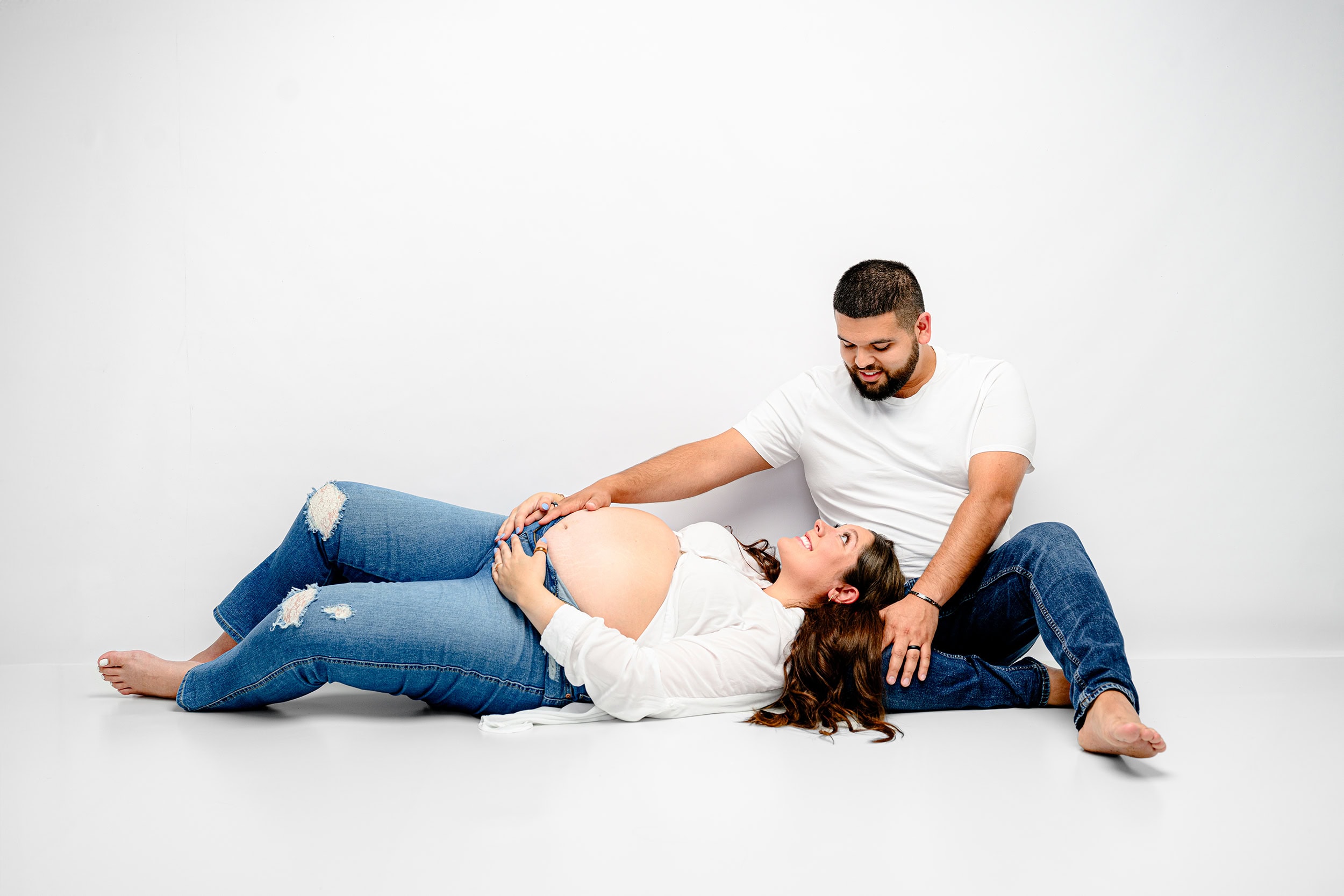 charlotte pregnant mother lays in her husbands lap wearing a open button white shirt and jeans with her belly exposed and her husband is wearing complimentary clothes while smiling down at her touching her belly