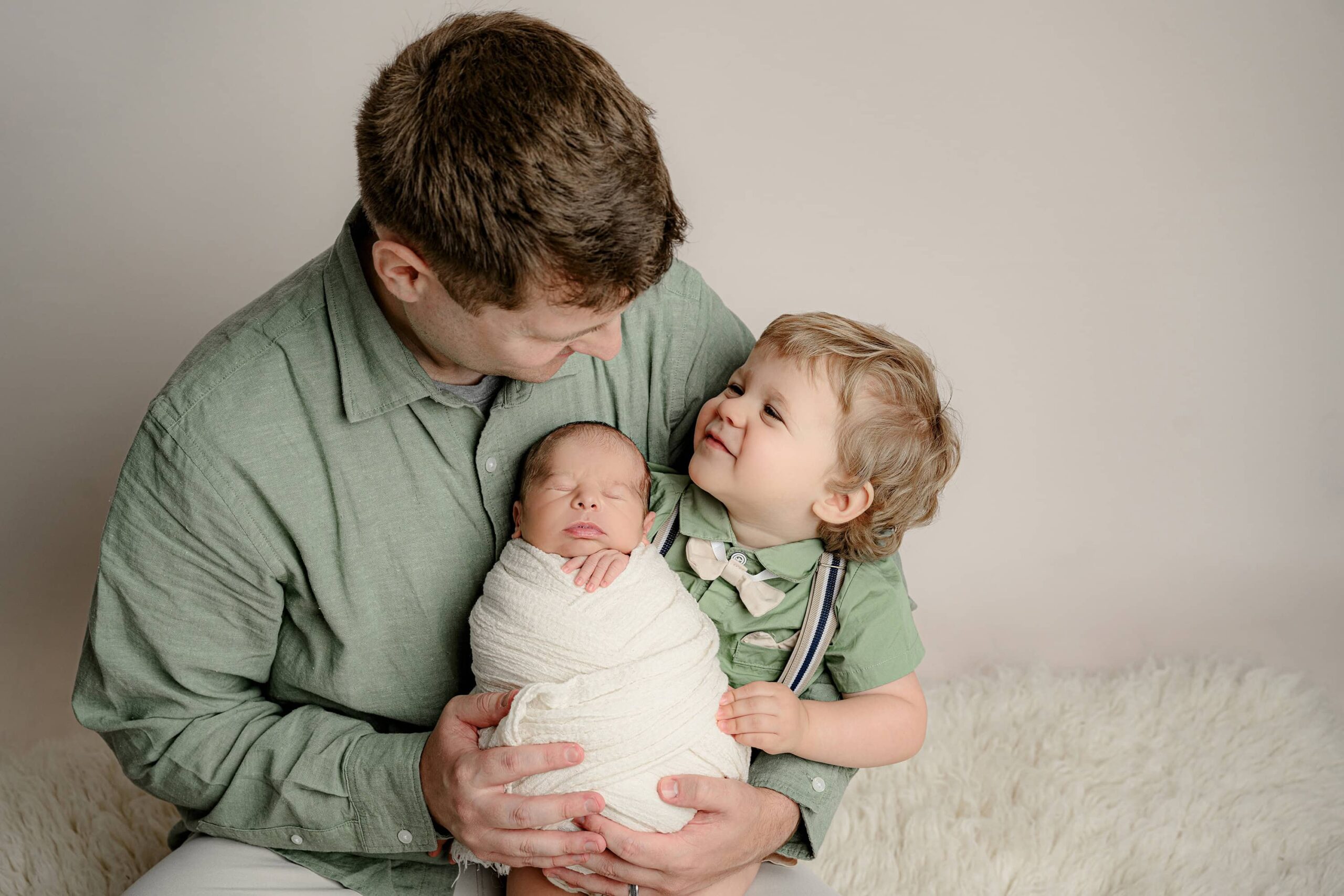 father holds his son and newborn in his lap smiling down at his toddler who is smiling up at him proudly holding his brother all wrapped in white