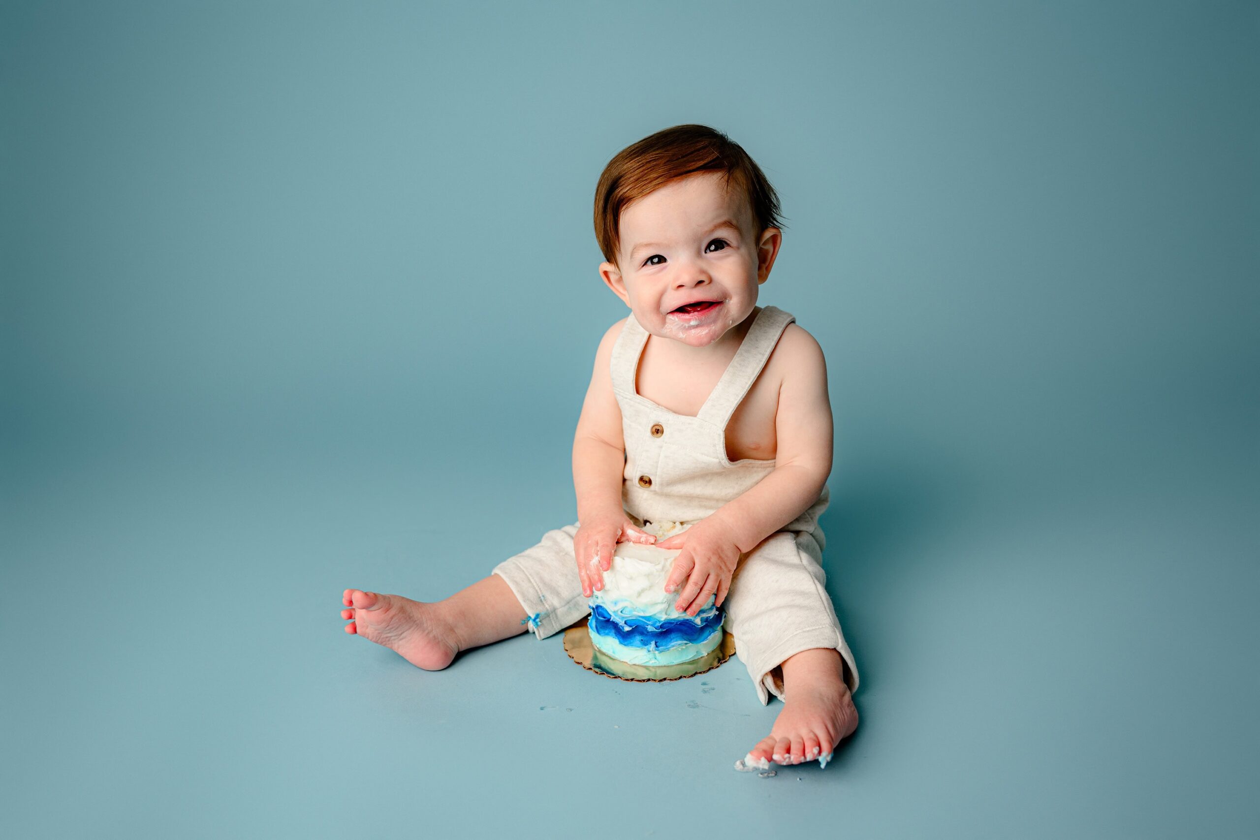 one year old little boy sits in a cream pair of overalls for his one year old milestone session on a blue background smashing his cake