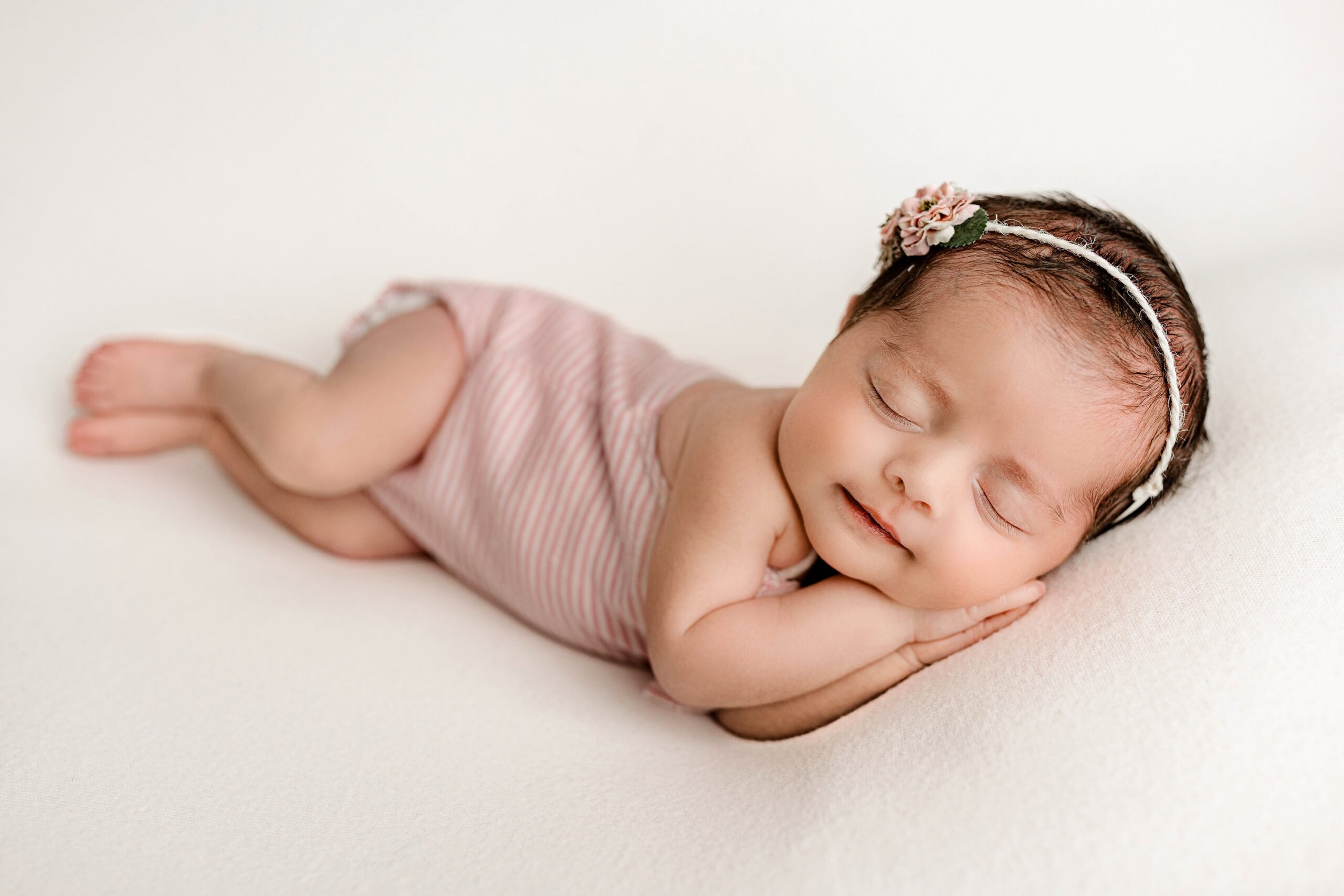 Charlotte newborn girl lays on her side with her hands under her cheek wearing a cream and white outfit and pink floral bow