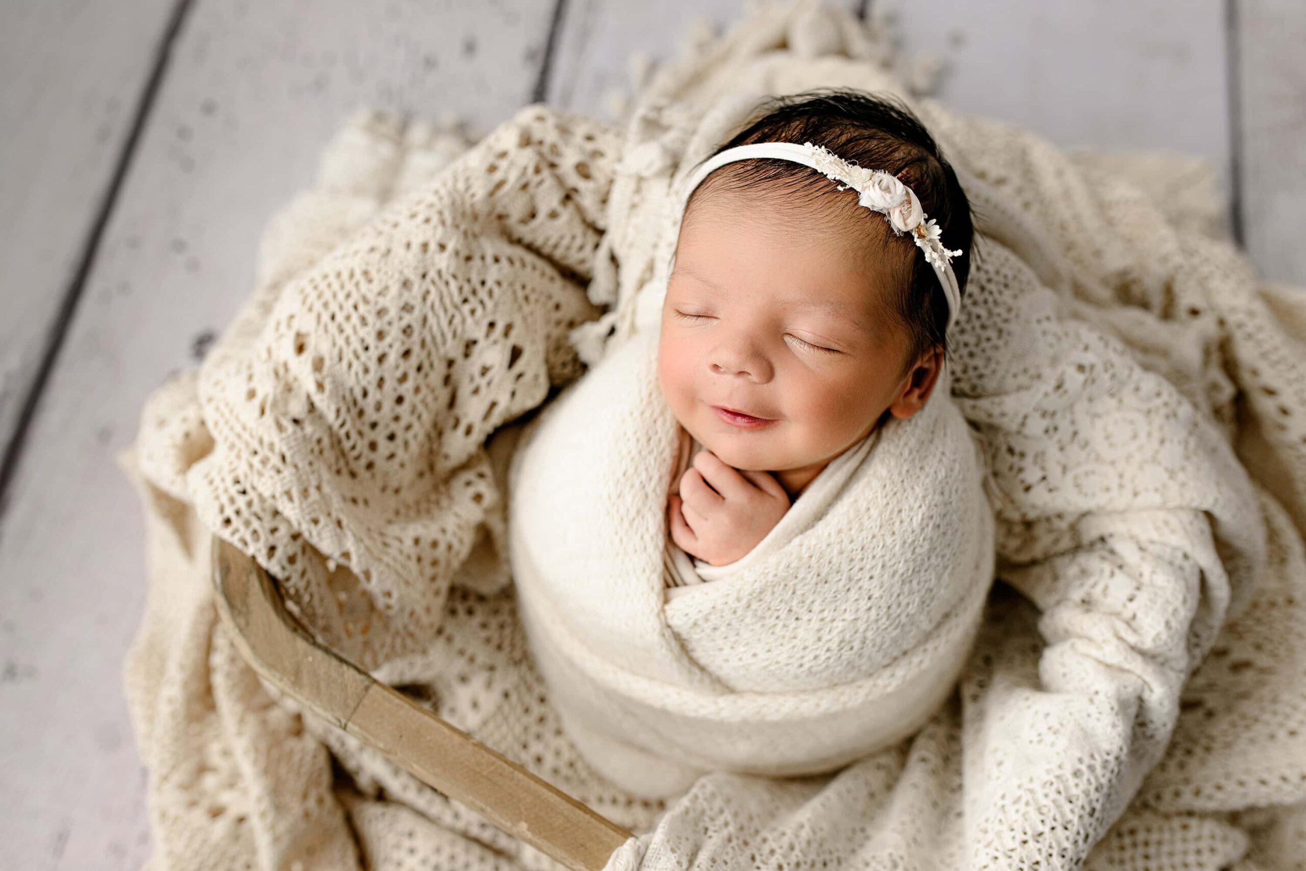 newborn baby girl from Charlotte, NC smiles while wrapped in a cream wrap wearing a cream bow posed in a basket with cream layers in it
